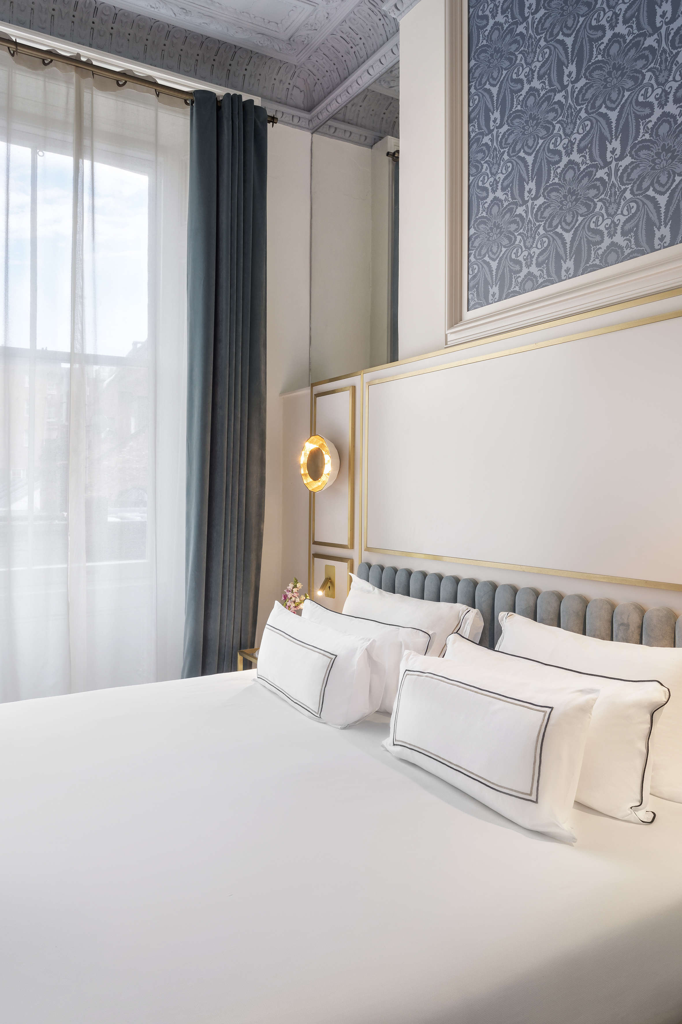 Elegant hotel bedroom: white bedding, gray tufted headboard, blue velvet curtains.