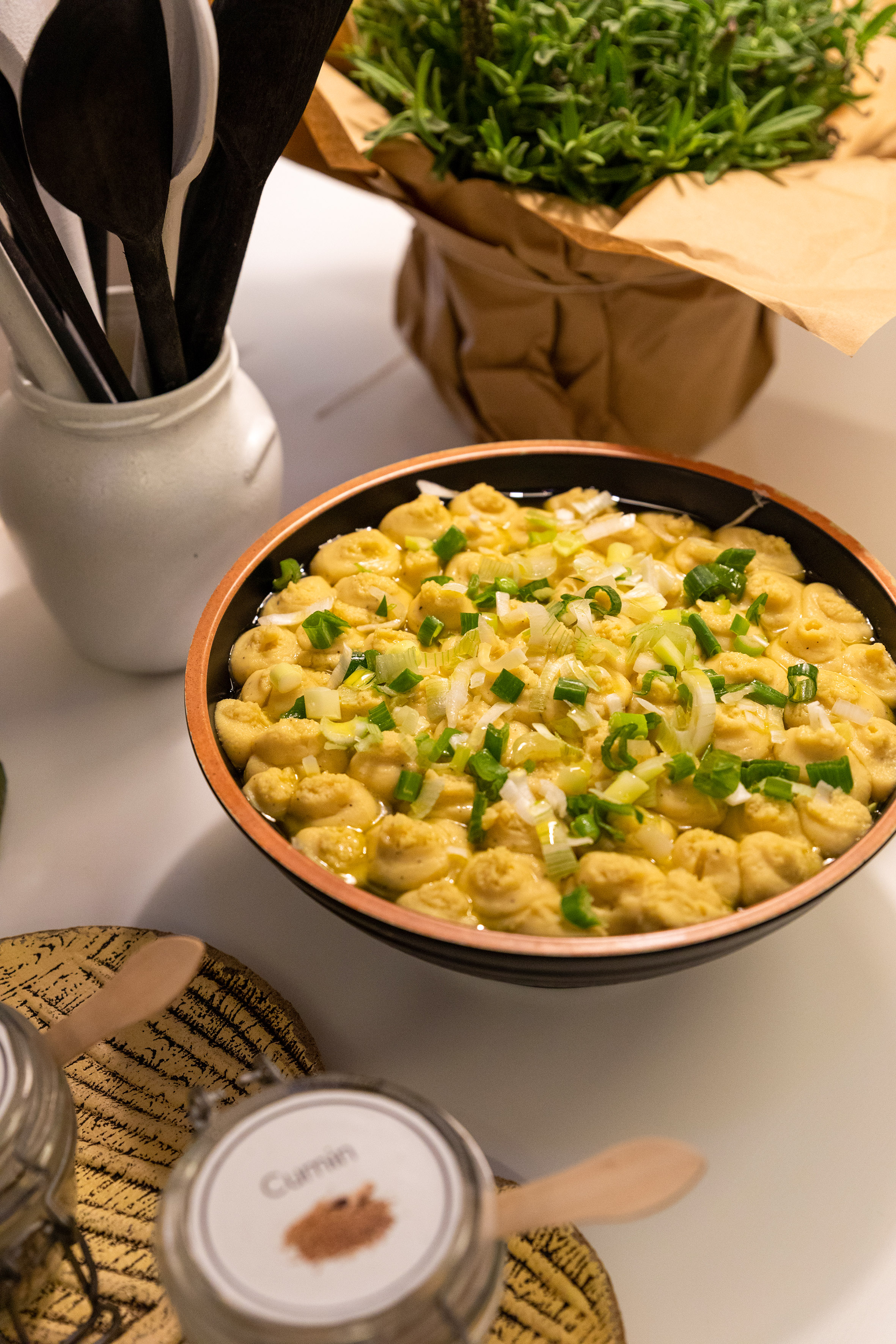 Dumplings with scallions in a bowl, kitchen utensils, cumin, and potted herbs.