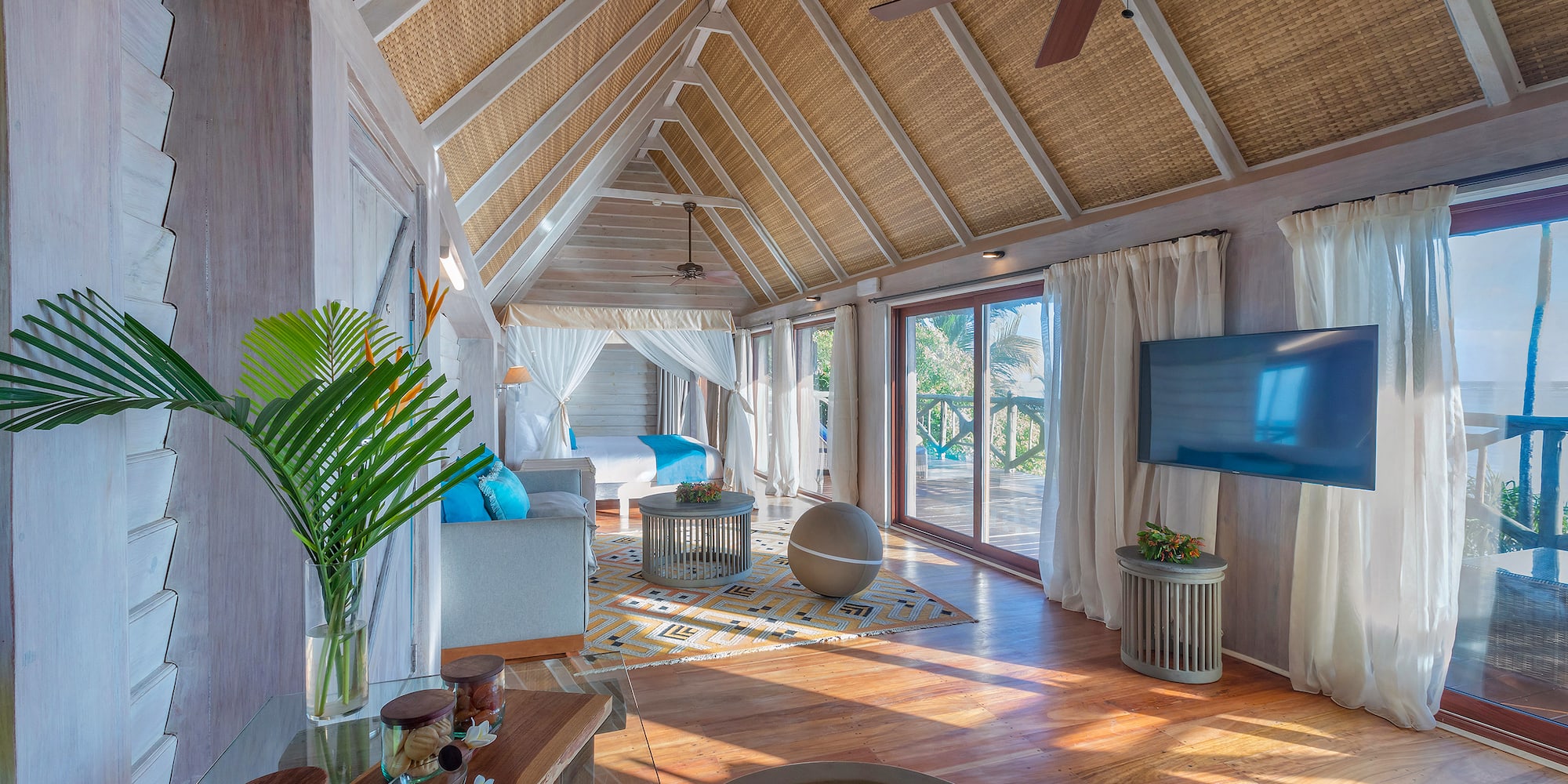 Spacious tropical resort suite with vaulted rattan ceiling, bed, and balcony.