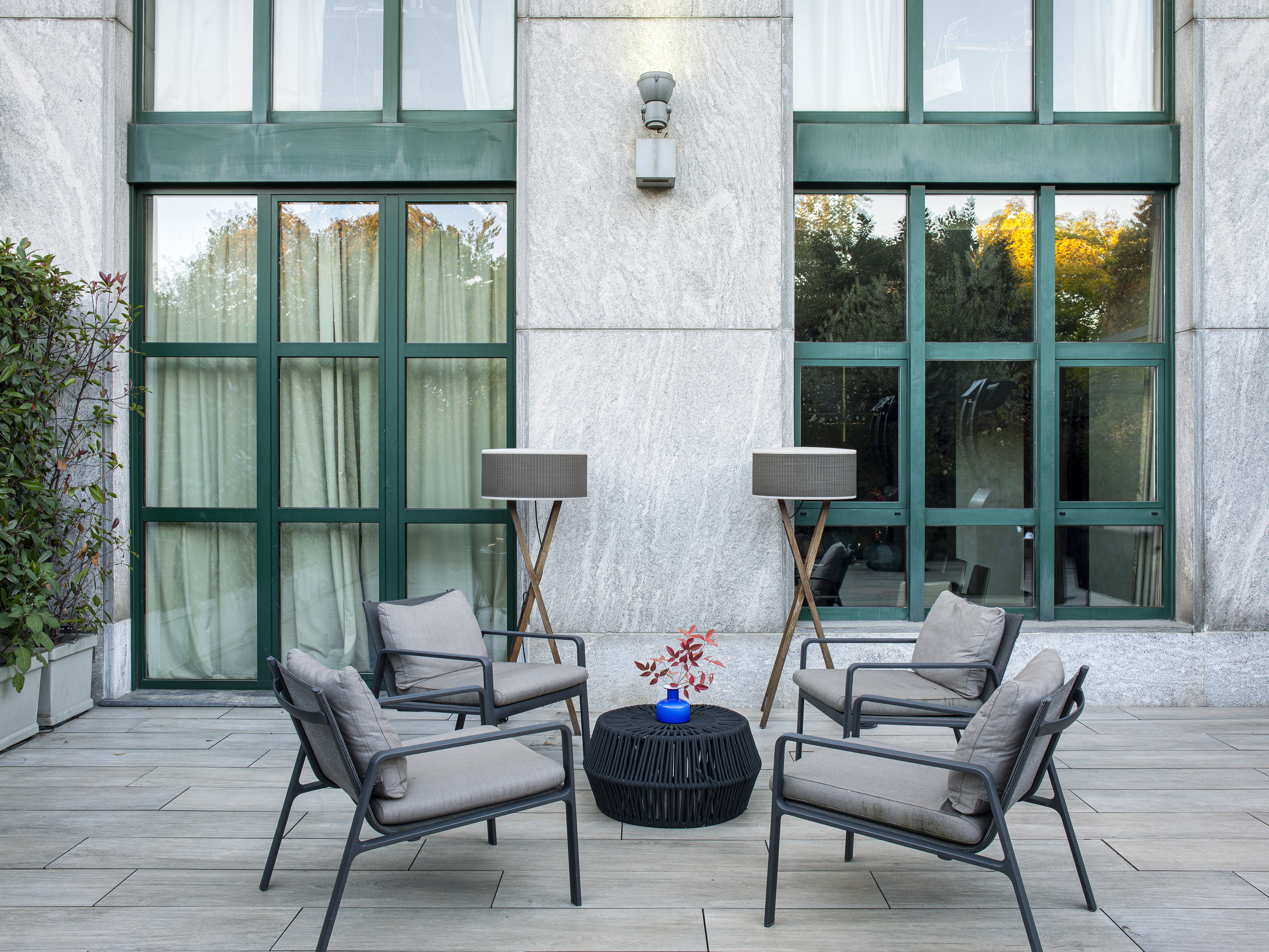 Modern patio lounge with grey chairs, black table, floor lamps, green windows.