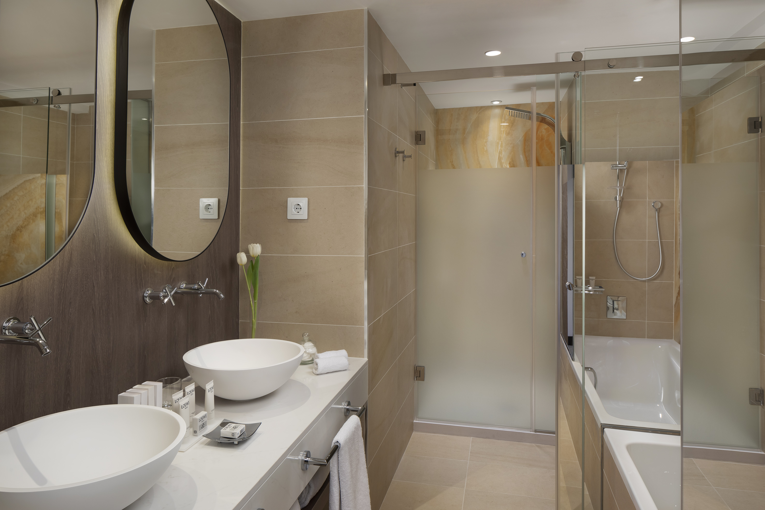 Modern beige bathroom with dual vessel sinks, backlit mirrors, shower, and tub.