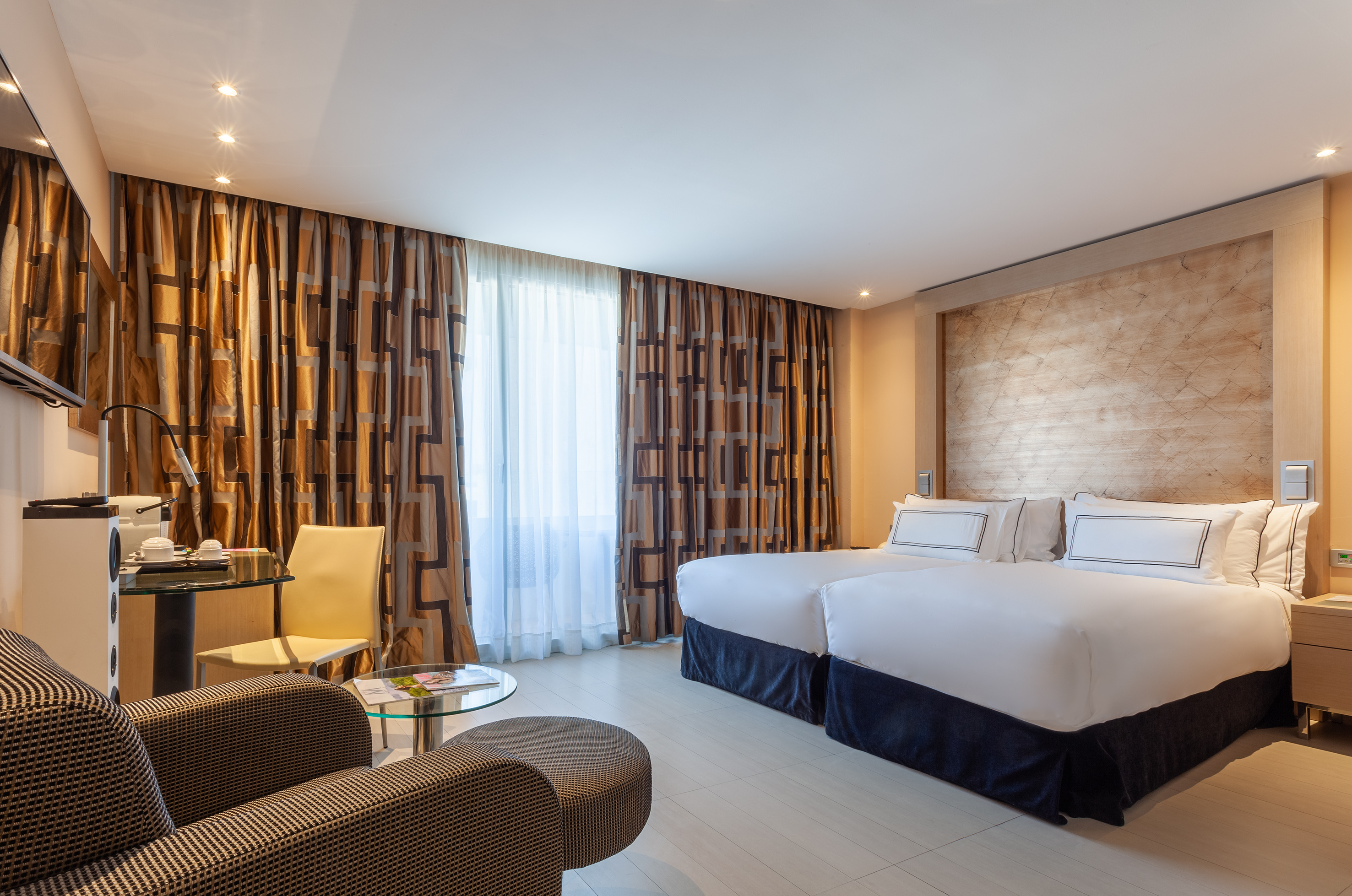 Modern hotel room with twin beds, patterned curtains, and seating area.