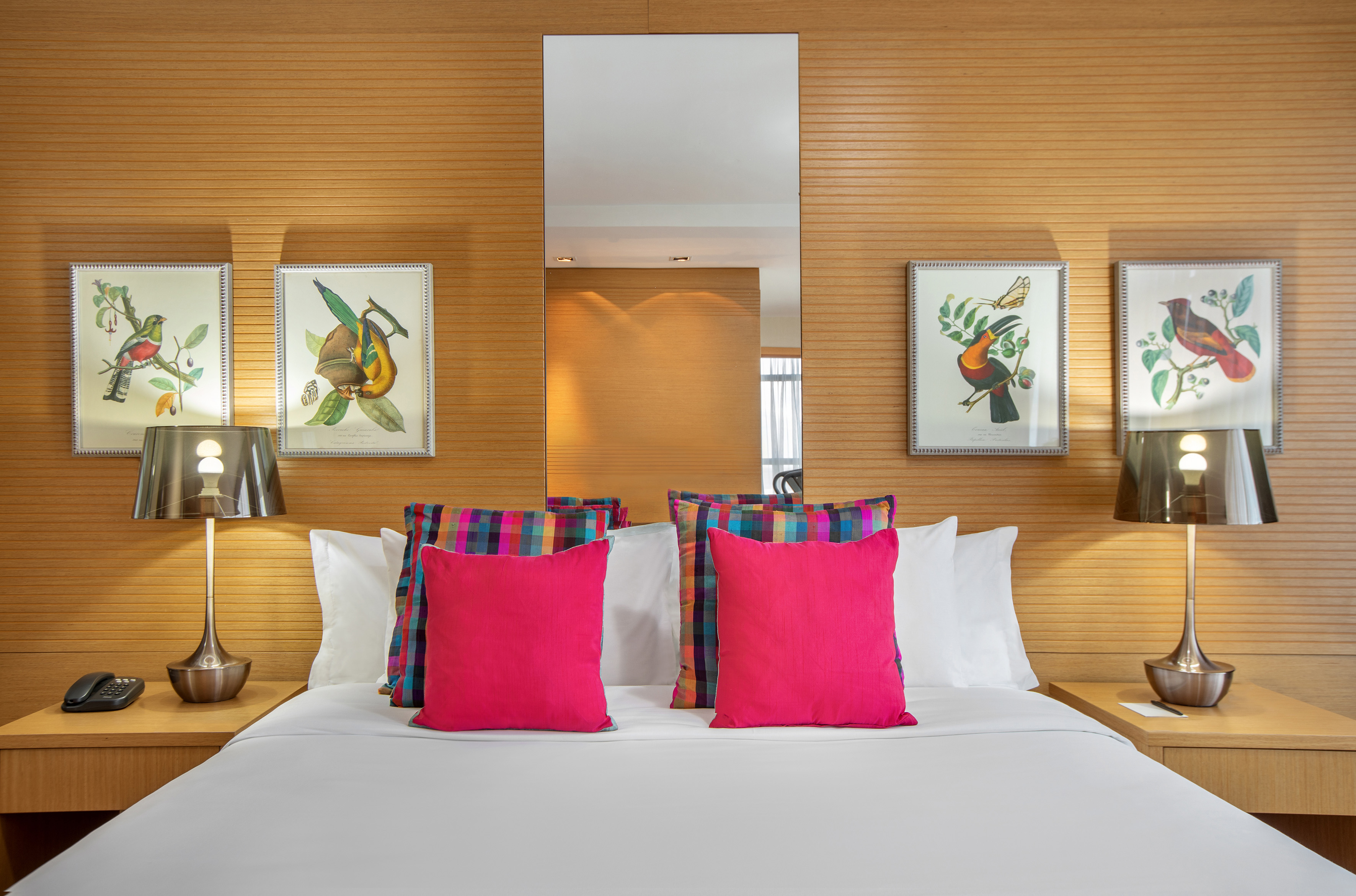 Hotel room bed with colorful pillows, wooden headboard, and framed bird art.