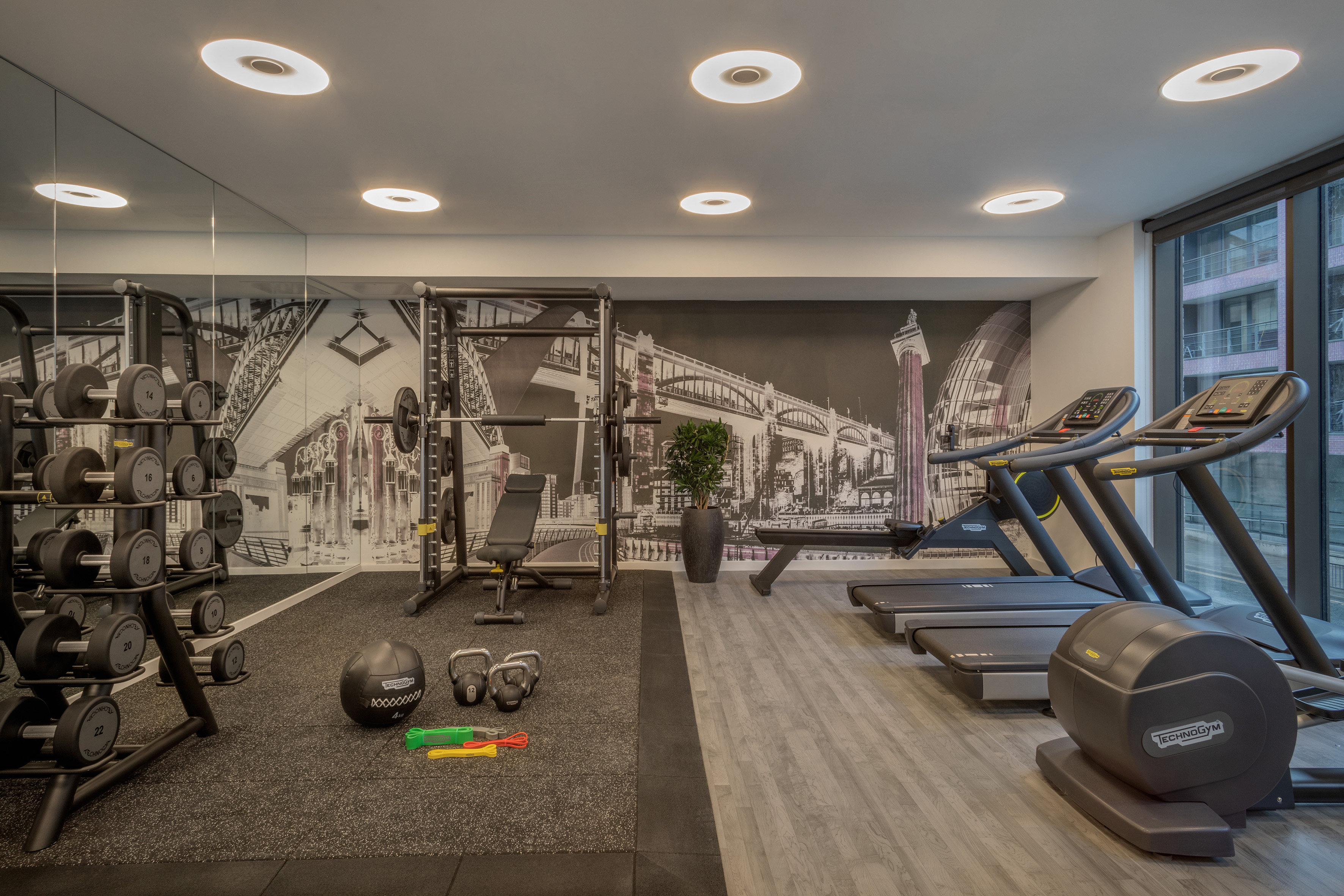 Modern fitness center with Technogym treadmills, weights, and cityscape mural.