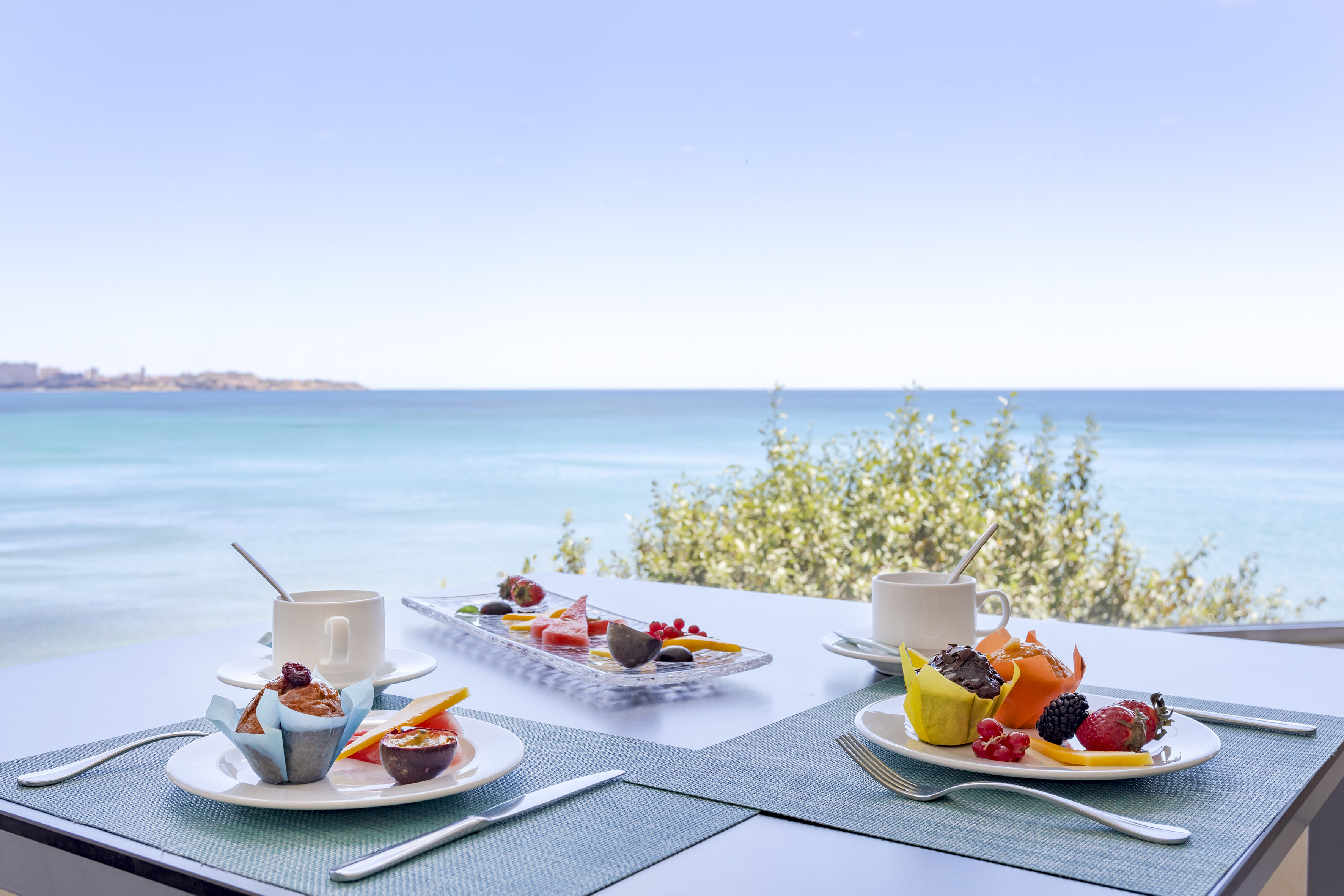 Outdoor breakfast with fresh fruit, muffins, and coffee, overlooking the sea.