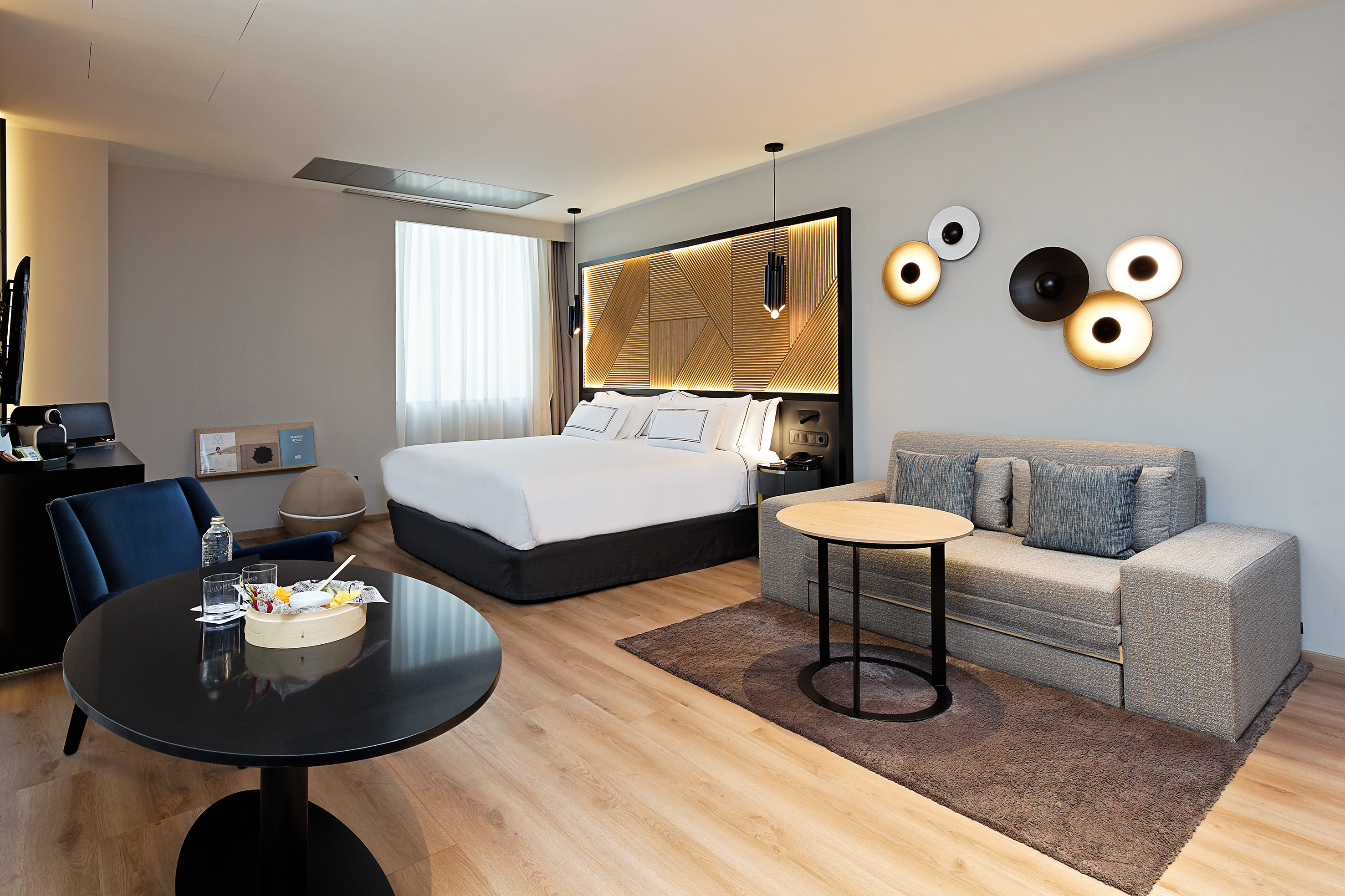 Modern hotel suite with king bed, sofa, coffee table, and contemporary lighting.