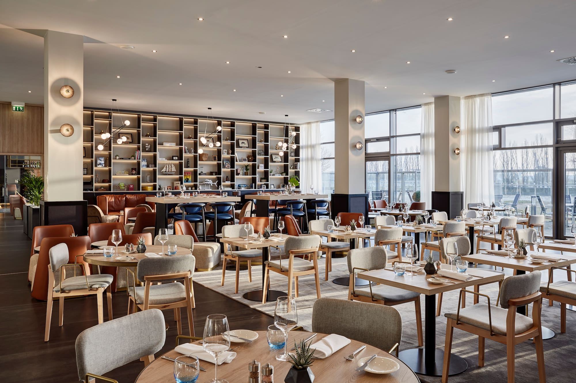 Modern restaurant dining area with set tables, bookshelves, and large windows.