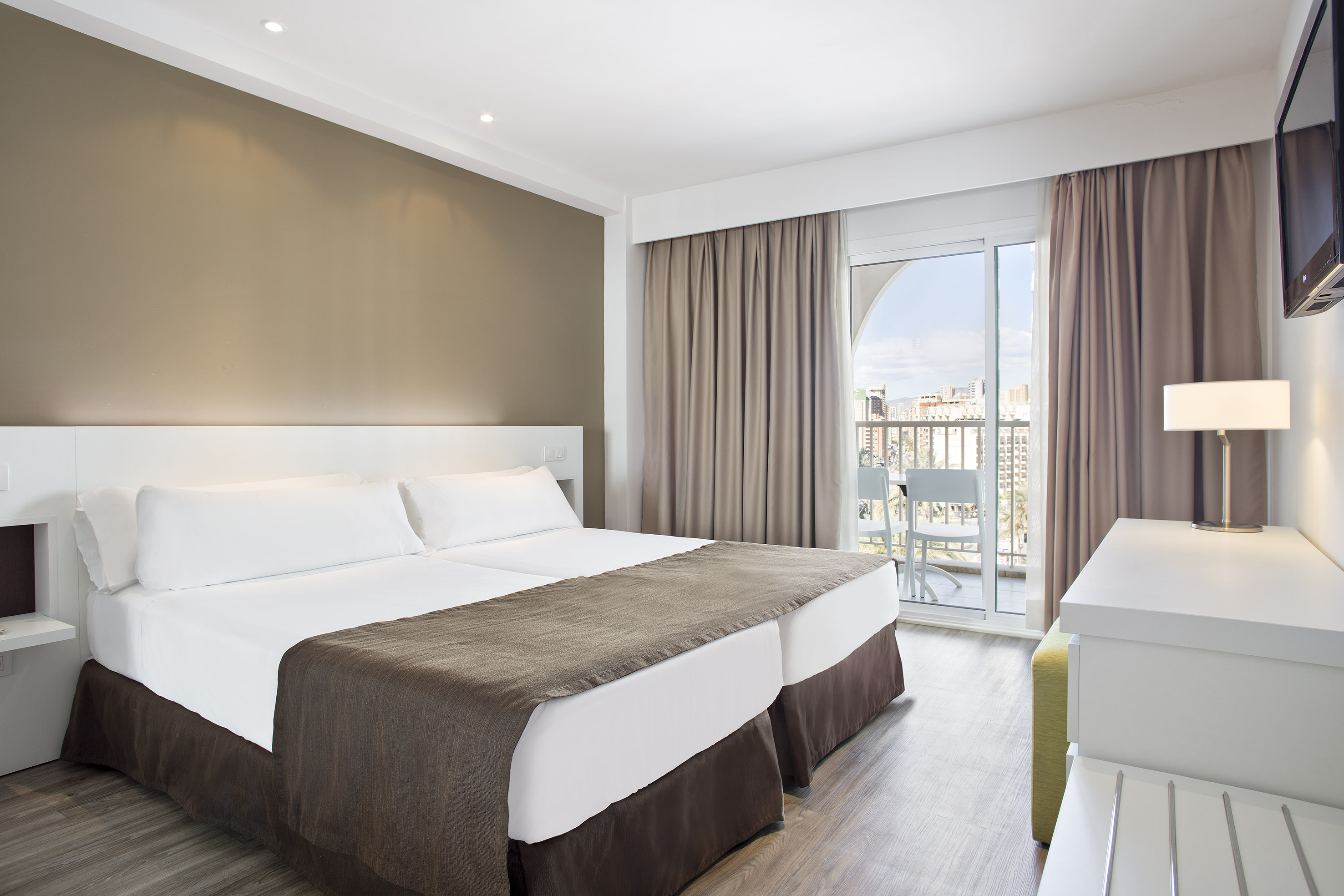 Modern hotel room with twin beds, white linens, and a balcony with city views.