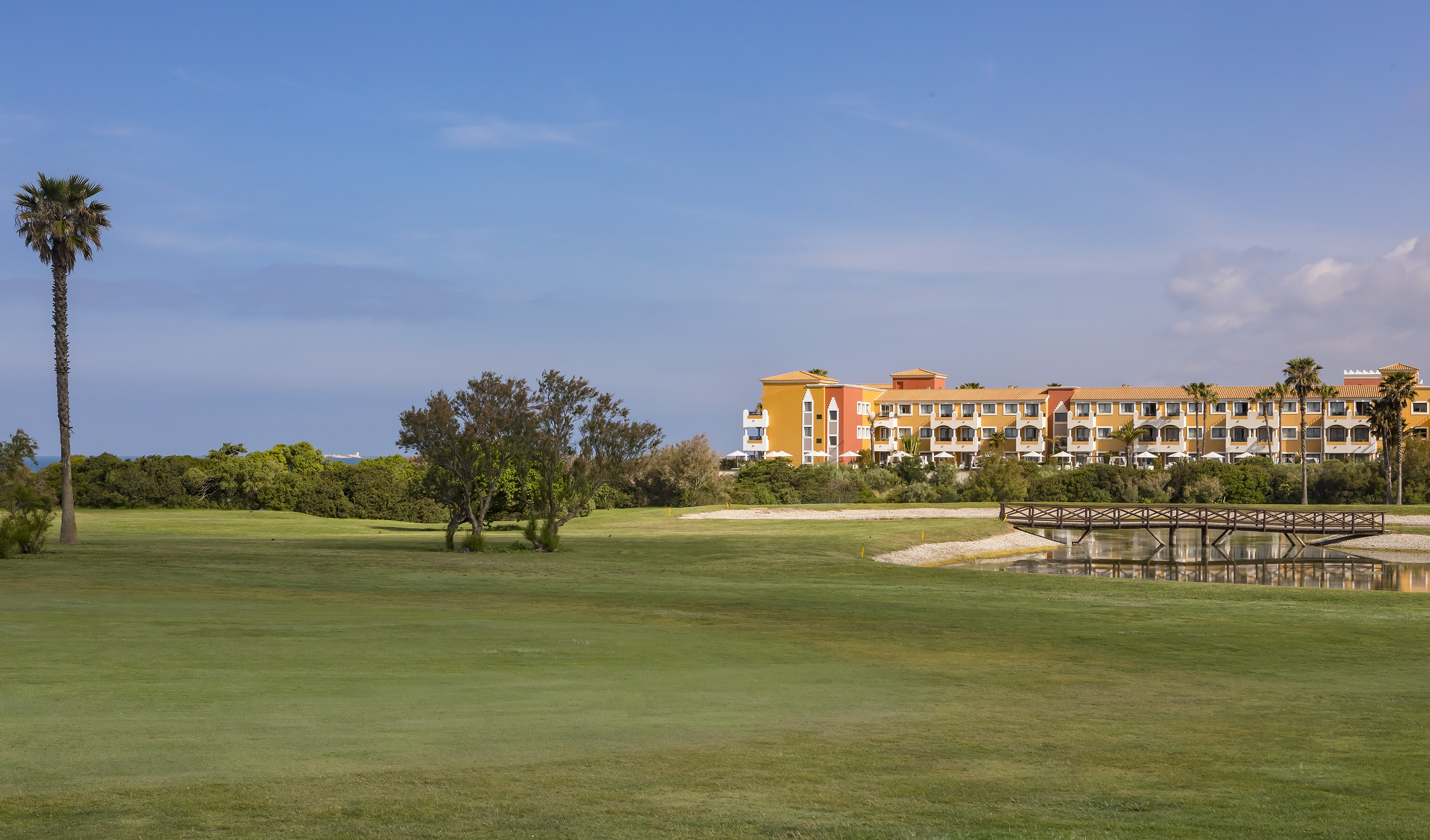 Colorful hotel and golf course with palm trees, bridge, and water feature.