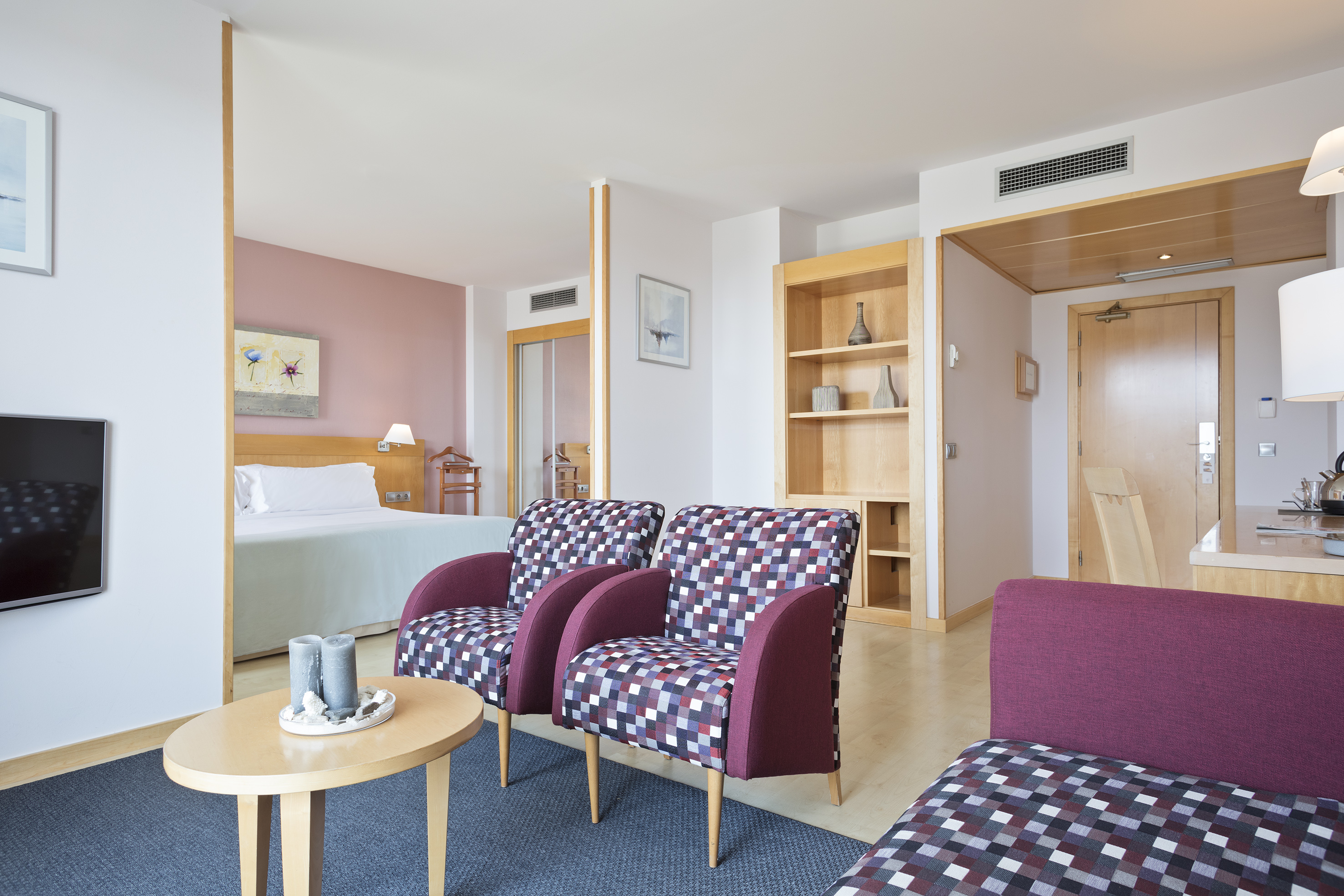 Modern hotel suite with living area, checkered armchairs, and separate bedroom.