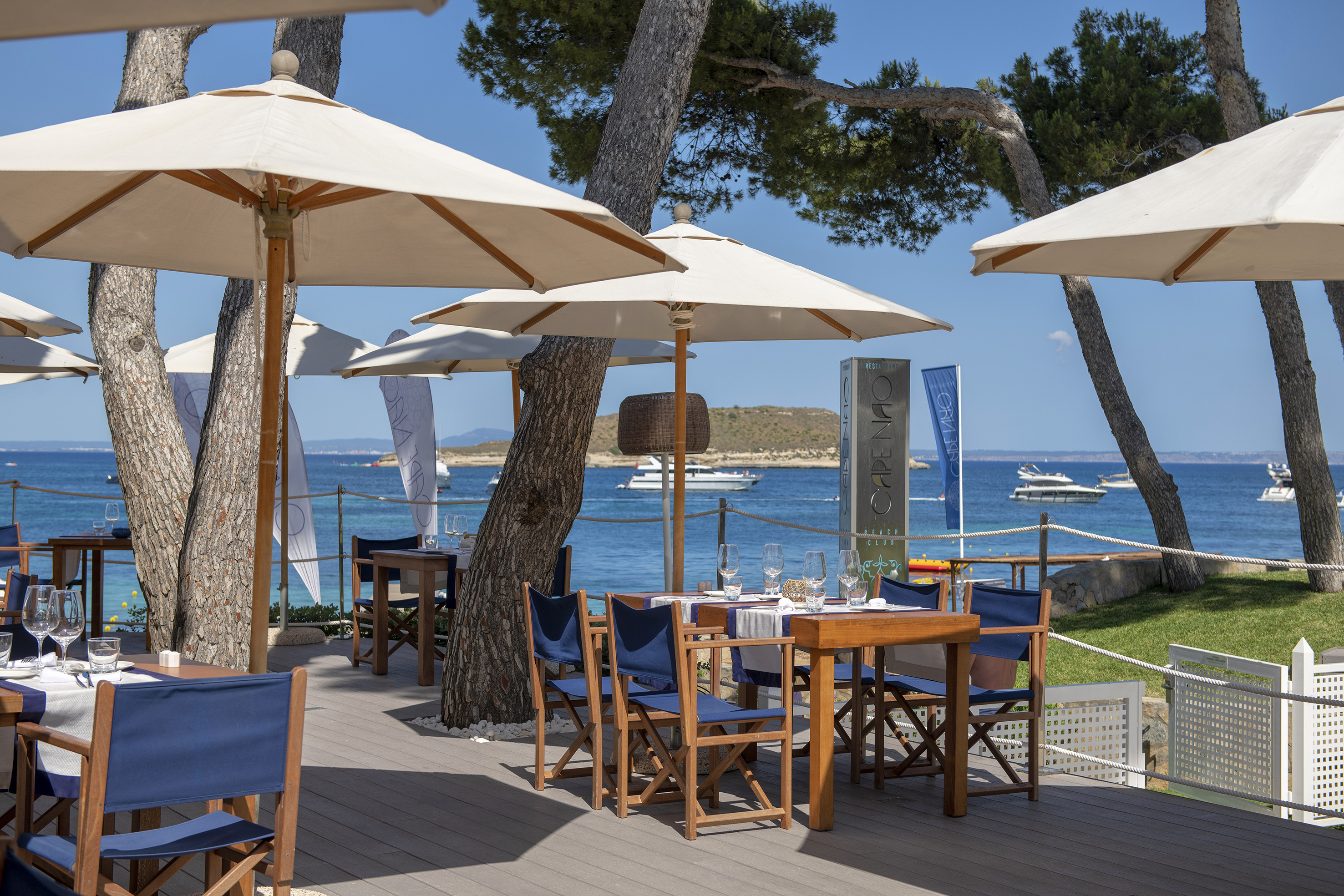 Beachfront dining deck with set tables, blue chairs, white umbrellas, and sea view.