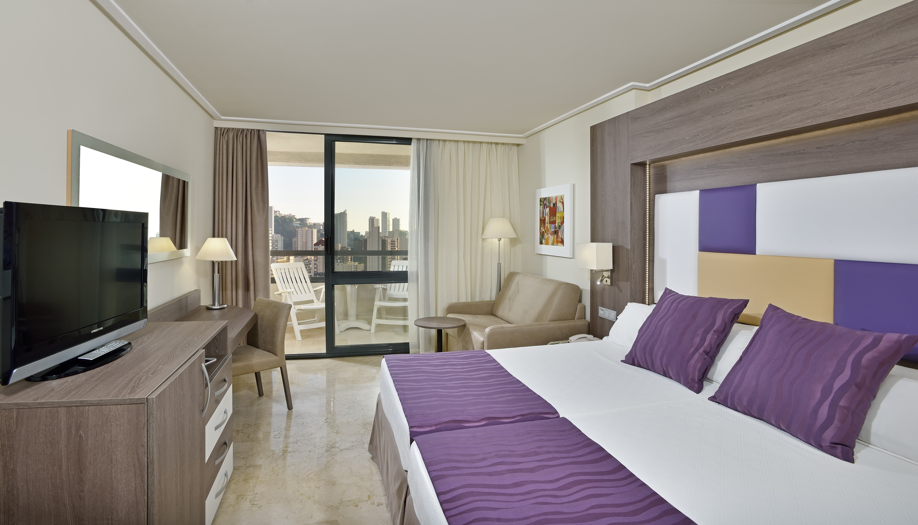 Modern hotel room with twin beds, purple bedding, balcony, city skyline view.