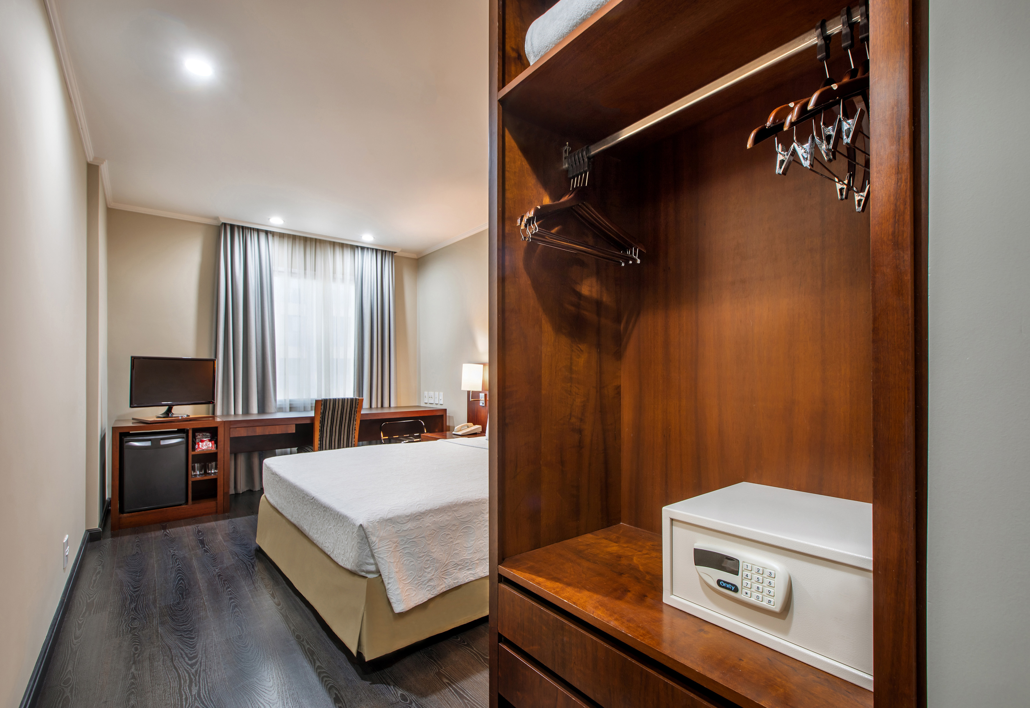Modern hotel guest room with open wooden closet, safe, bed, and desk.