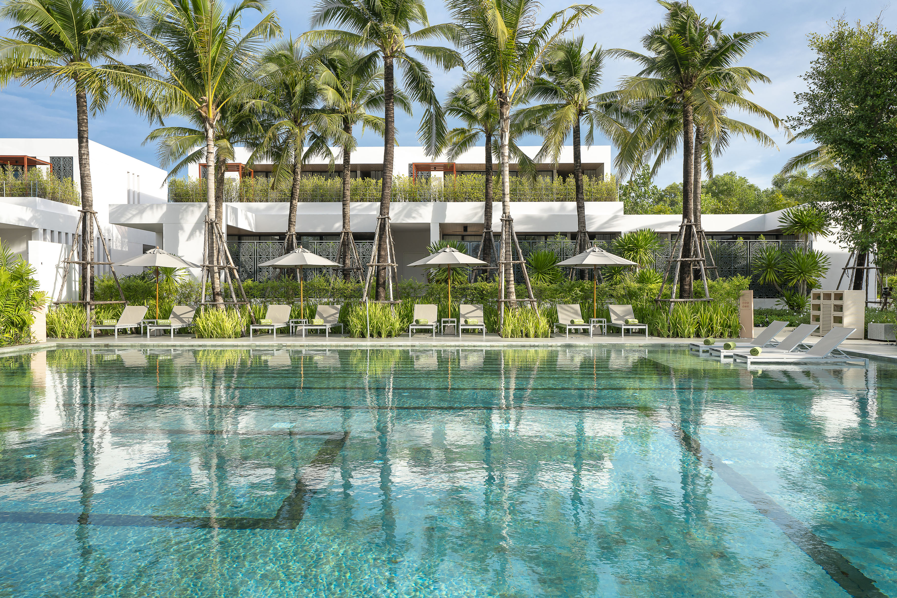 Modern resort swimming pool with palm trees, lounge chairs, and white buildings.