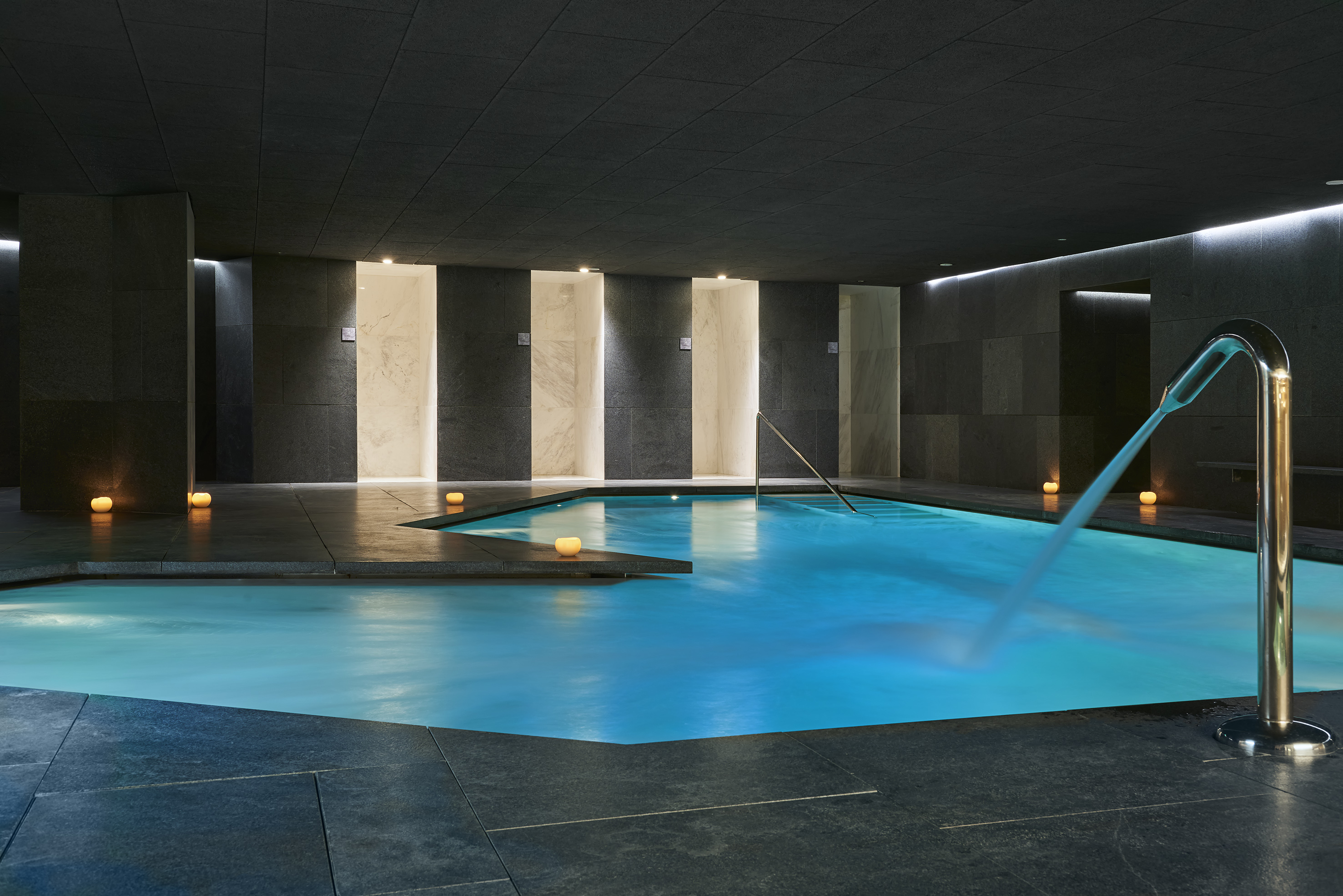 Dark luxury spa pool with blue water, waterfall, candlelit niches, and stone walls.