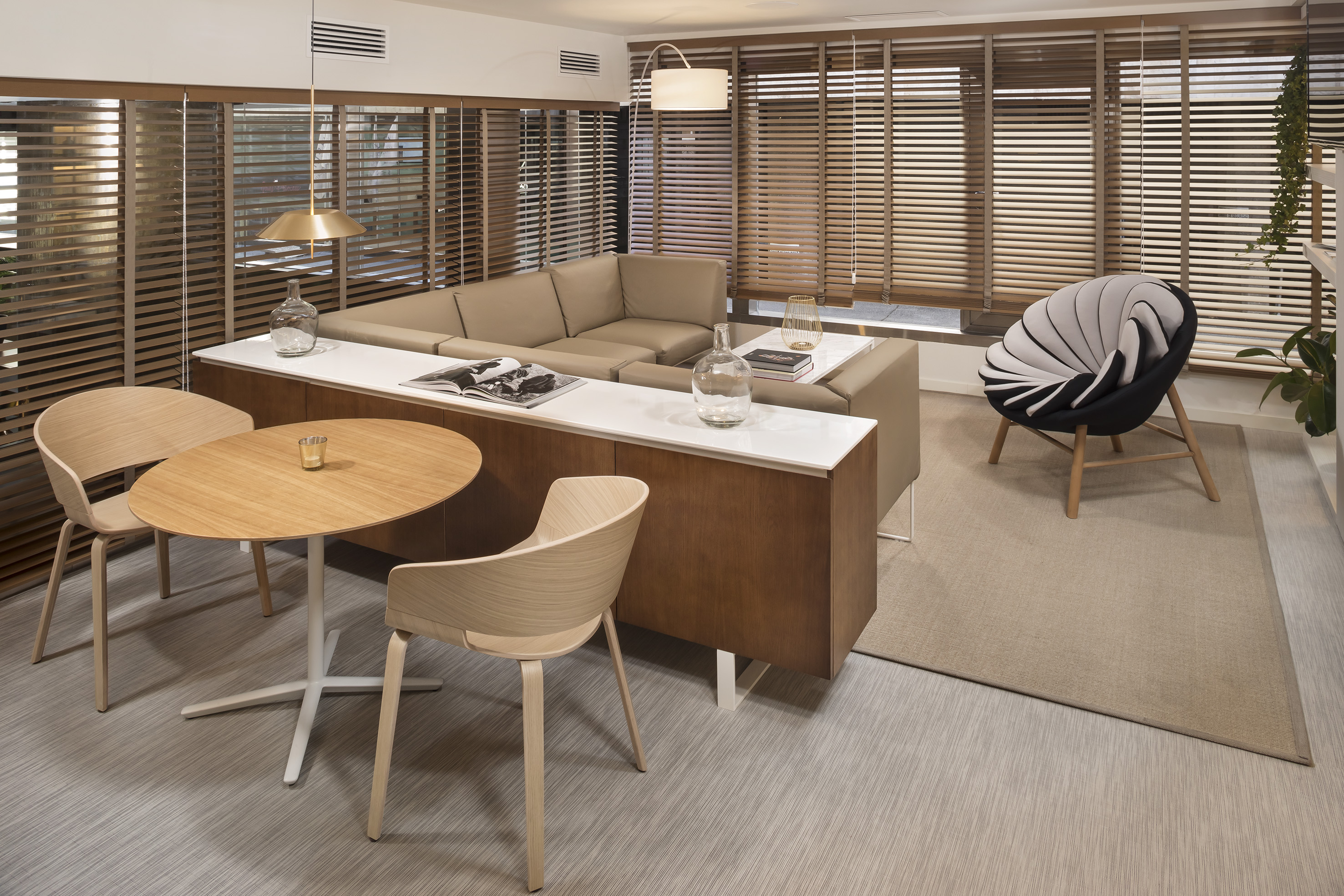 Modern office lounge with beige sofa, wooden table, accent chair, and window blinds.