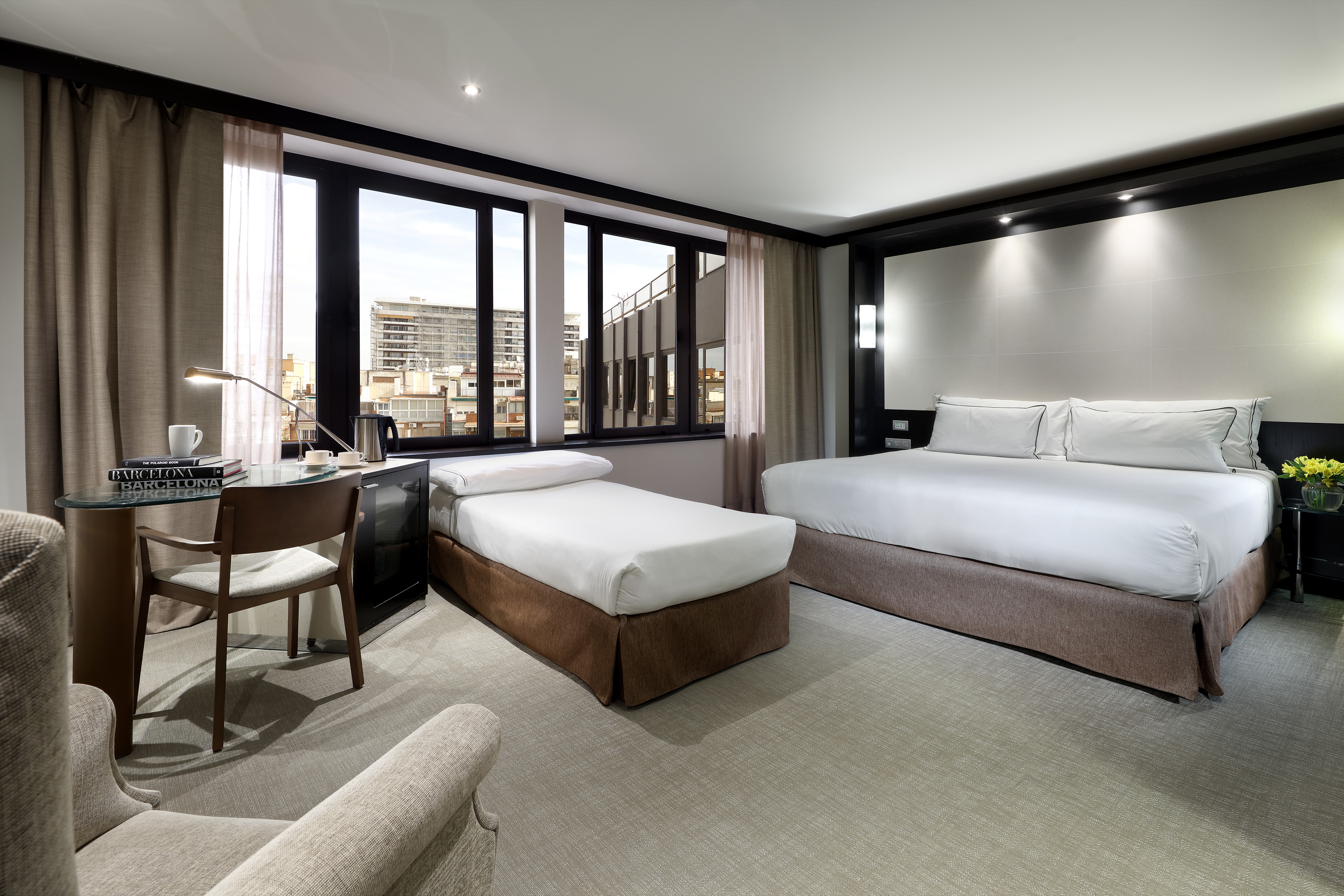 Modern hotel room with king, single bed, desk, and city view.