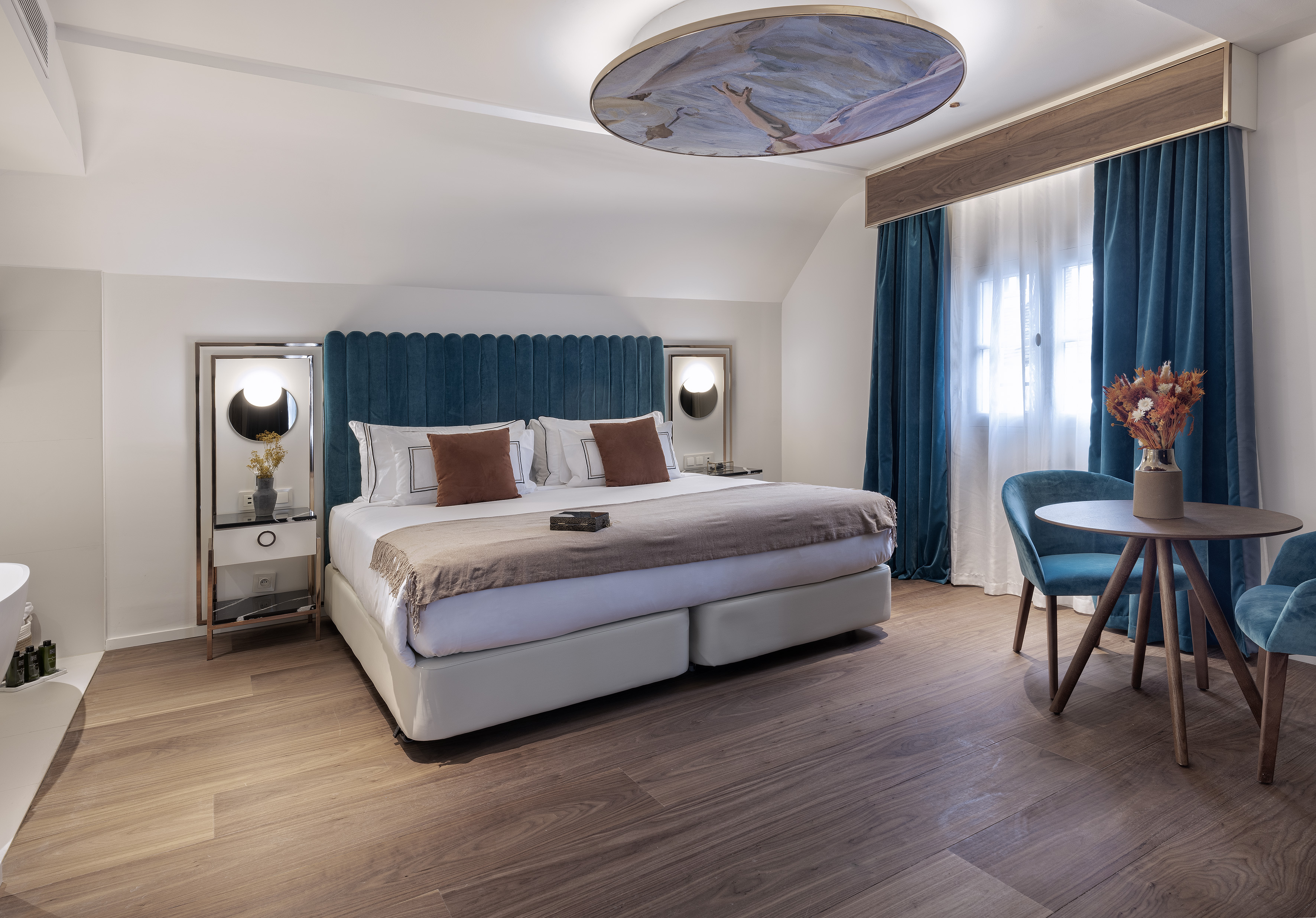 Modern hotel bedroom with blue velvet bed, artistic ceiling, and wood floor.
