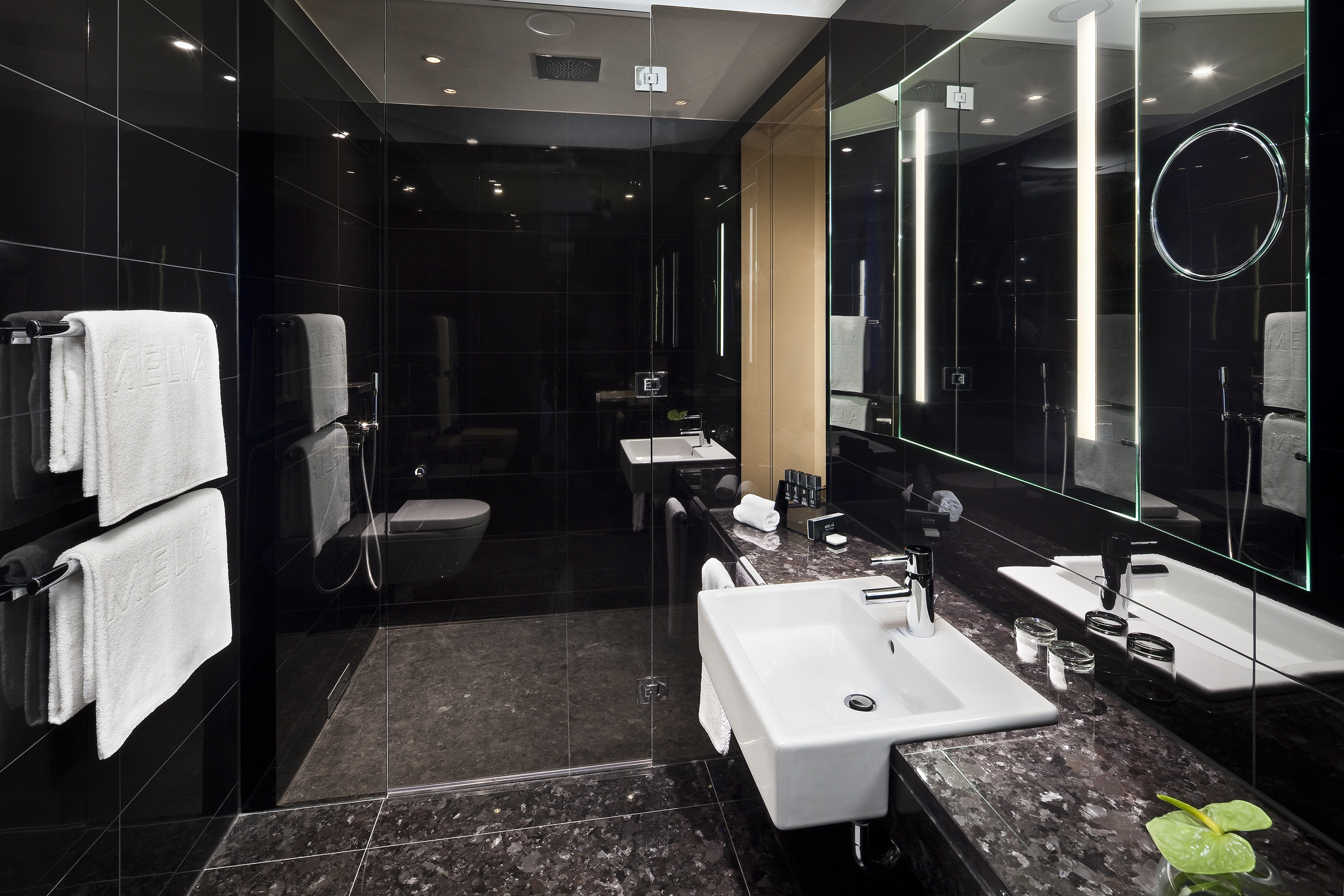 Modern bathroom with black tiles, granite vanity, white sinks, and illuminated mirrors.