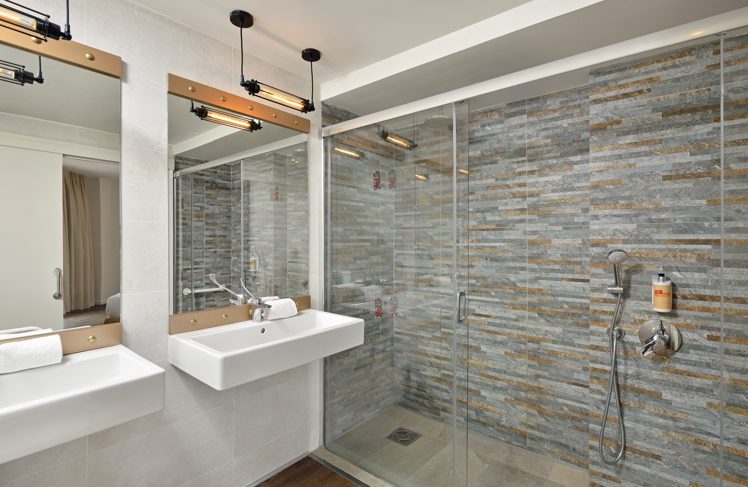 Modern hotel bathroom with double vanity, walk-in shower, and industrial lighting.