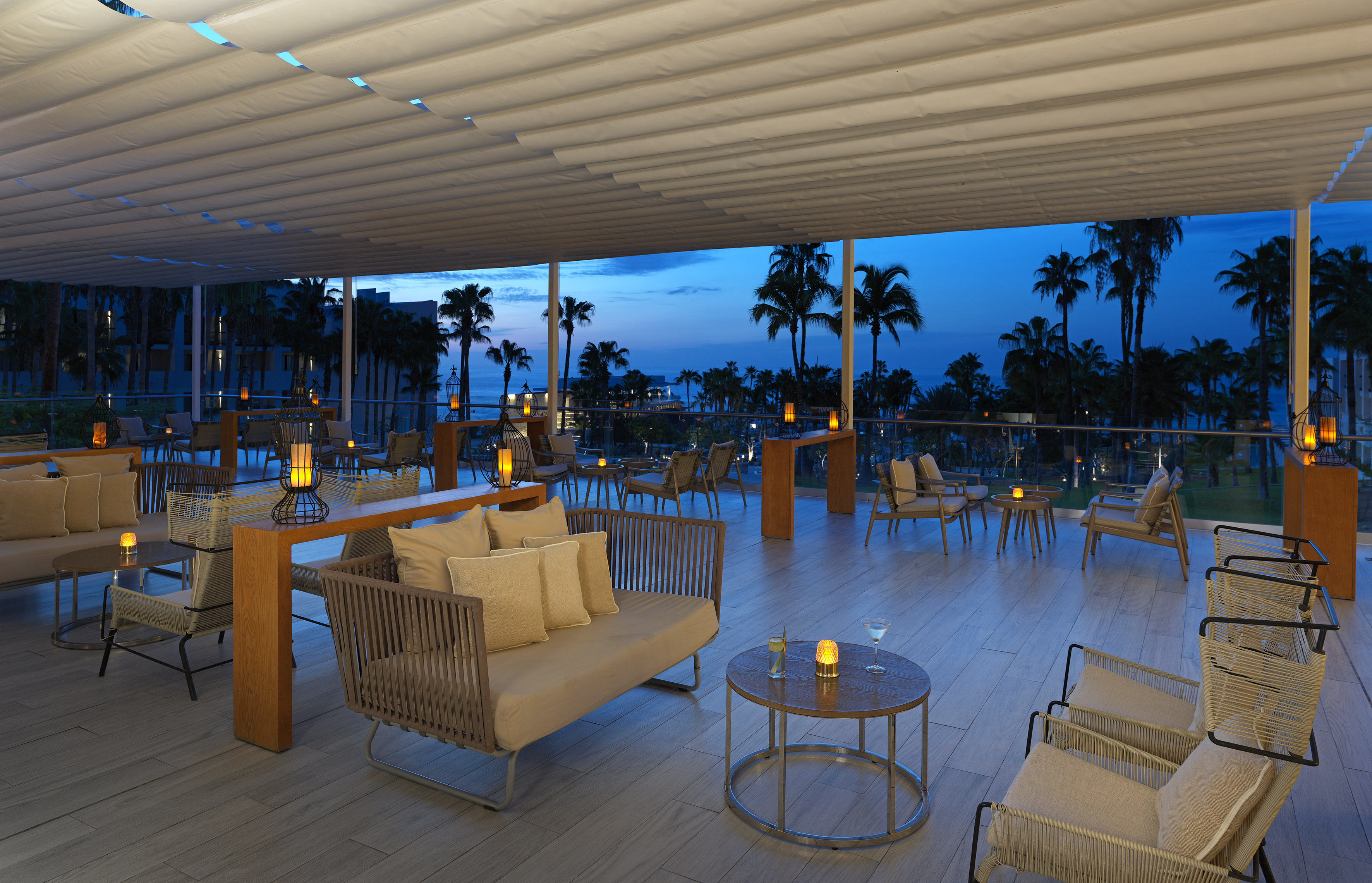 Evening resort lounge patio with ocean view, palm trees, and ambient lighting.
