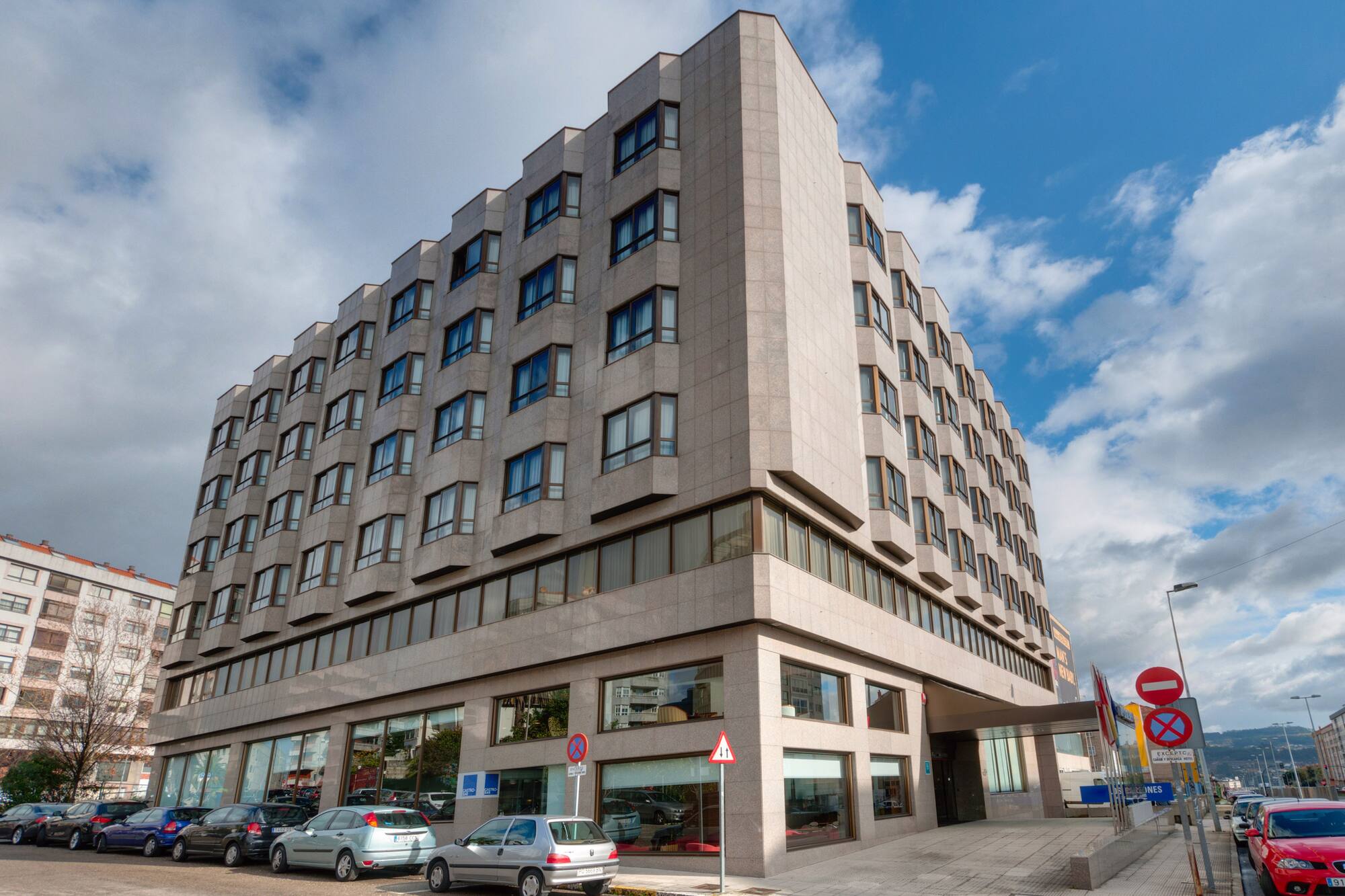 Modern hotel building facade with light stone, many windows, and parked cars.