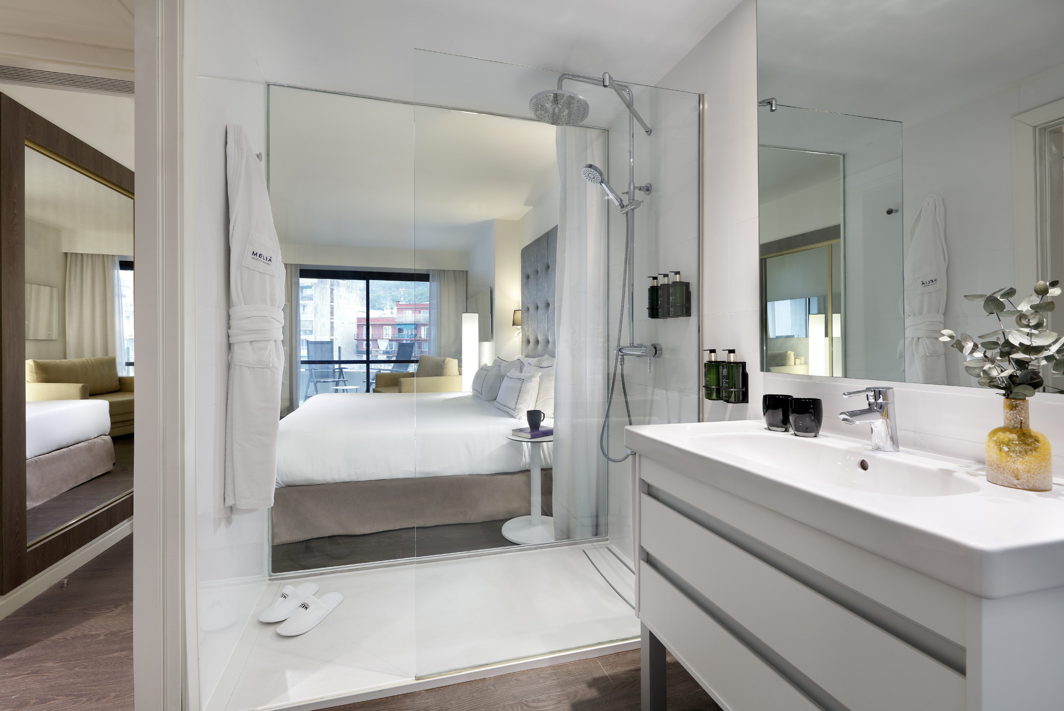 Open-plan hotel suite with glass shower, modern vanity, and bedroom.
