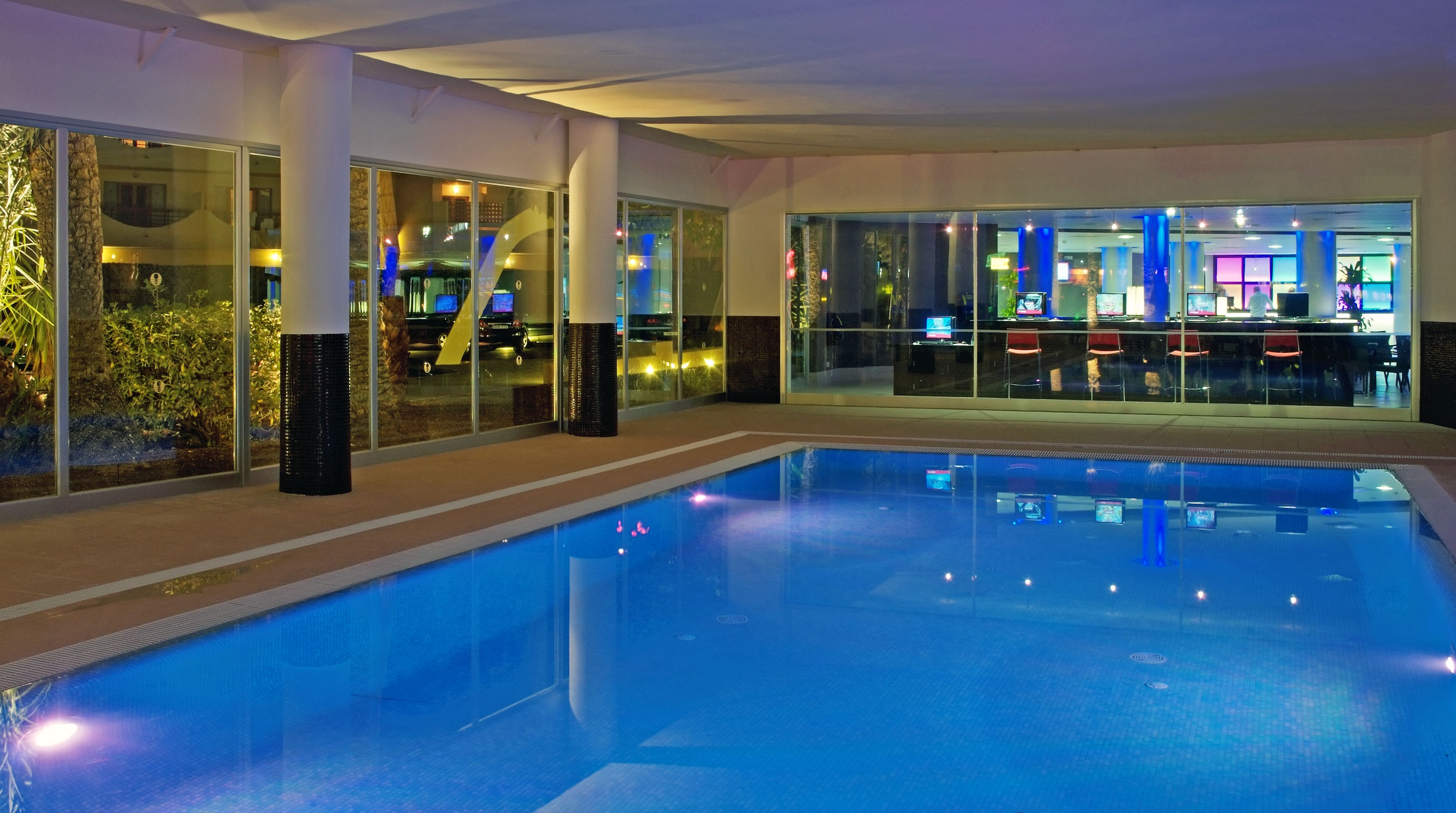 Indoor hotel pool illuminated at night, next to a computer lounge.