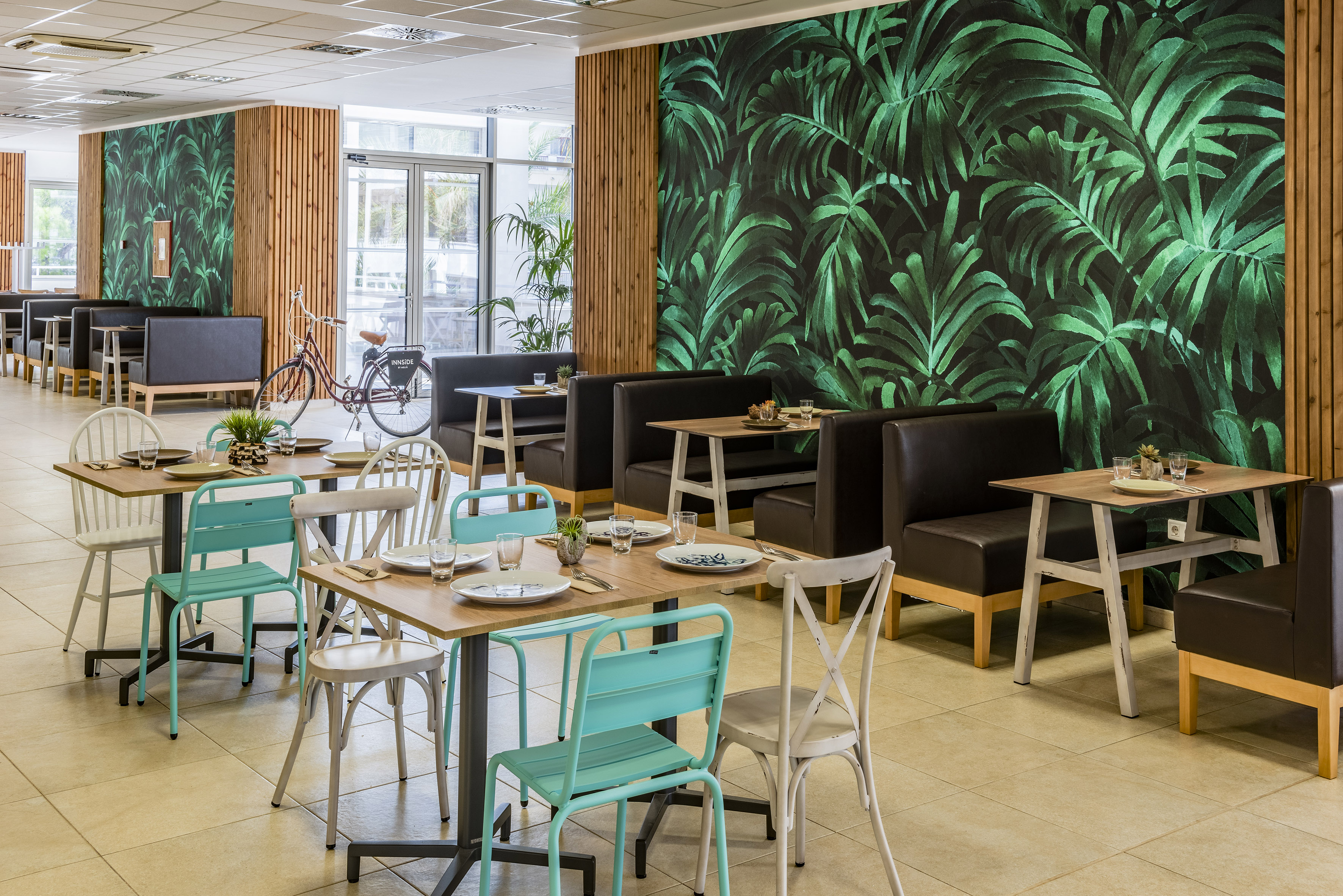 Modern restaurant interior with tropical leaf wallpaper, booth seating, and tables.