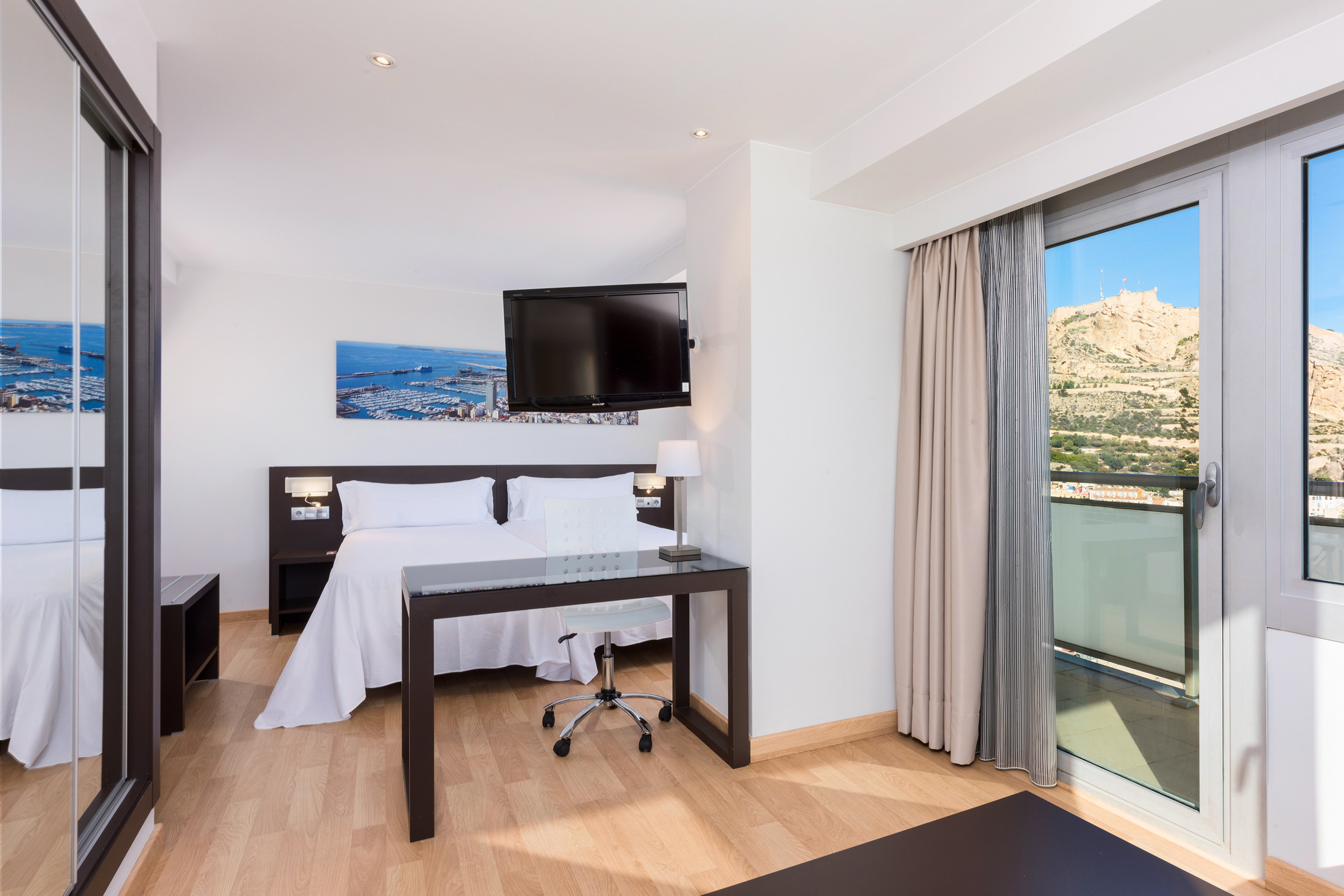 Modern hotel room with balcony, desk, TV, and Alicante castle mountain view.