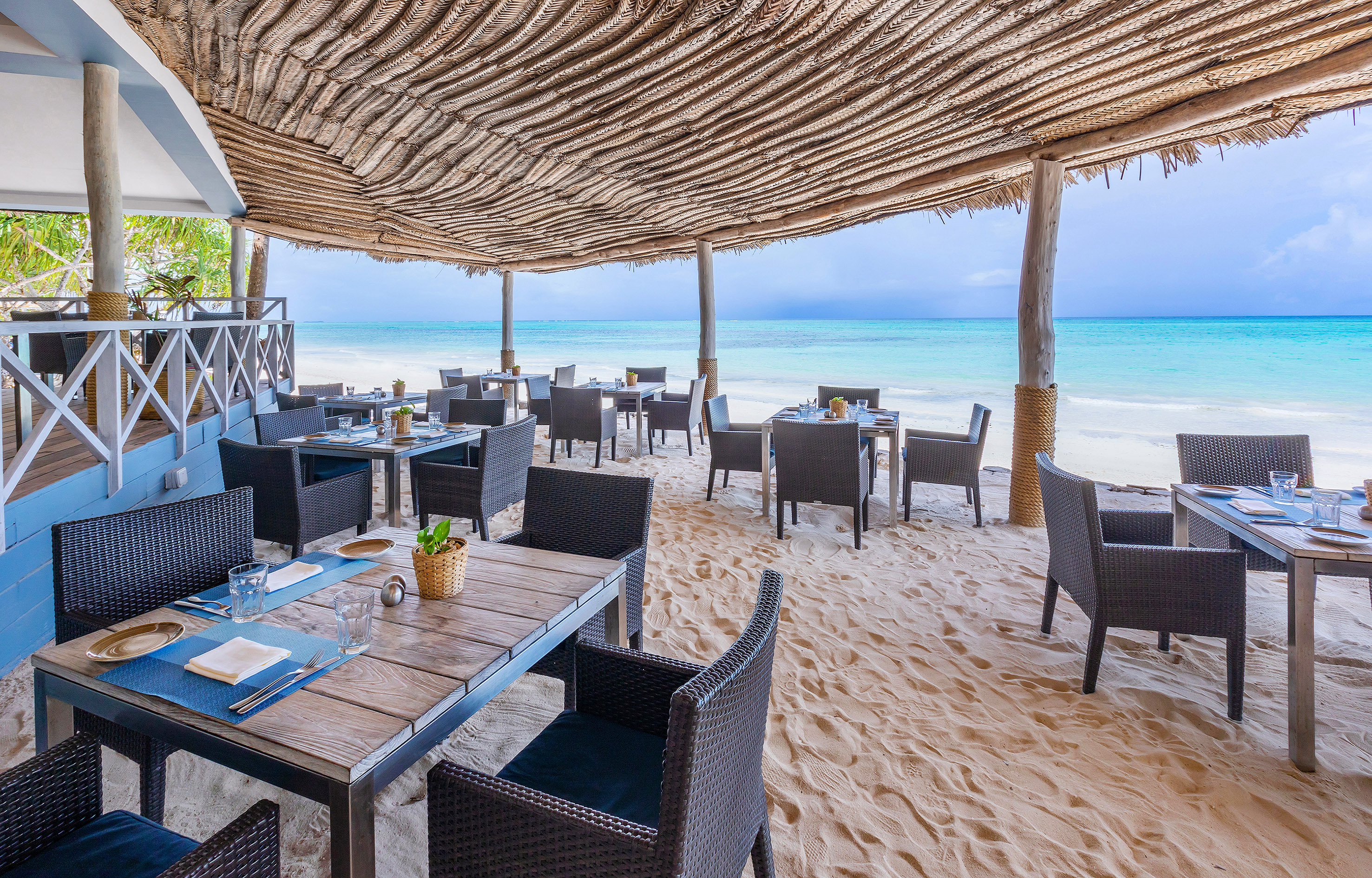 Beachfront restaurant with thatched roof, tables on sand, and turquoise ocean.