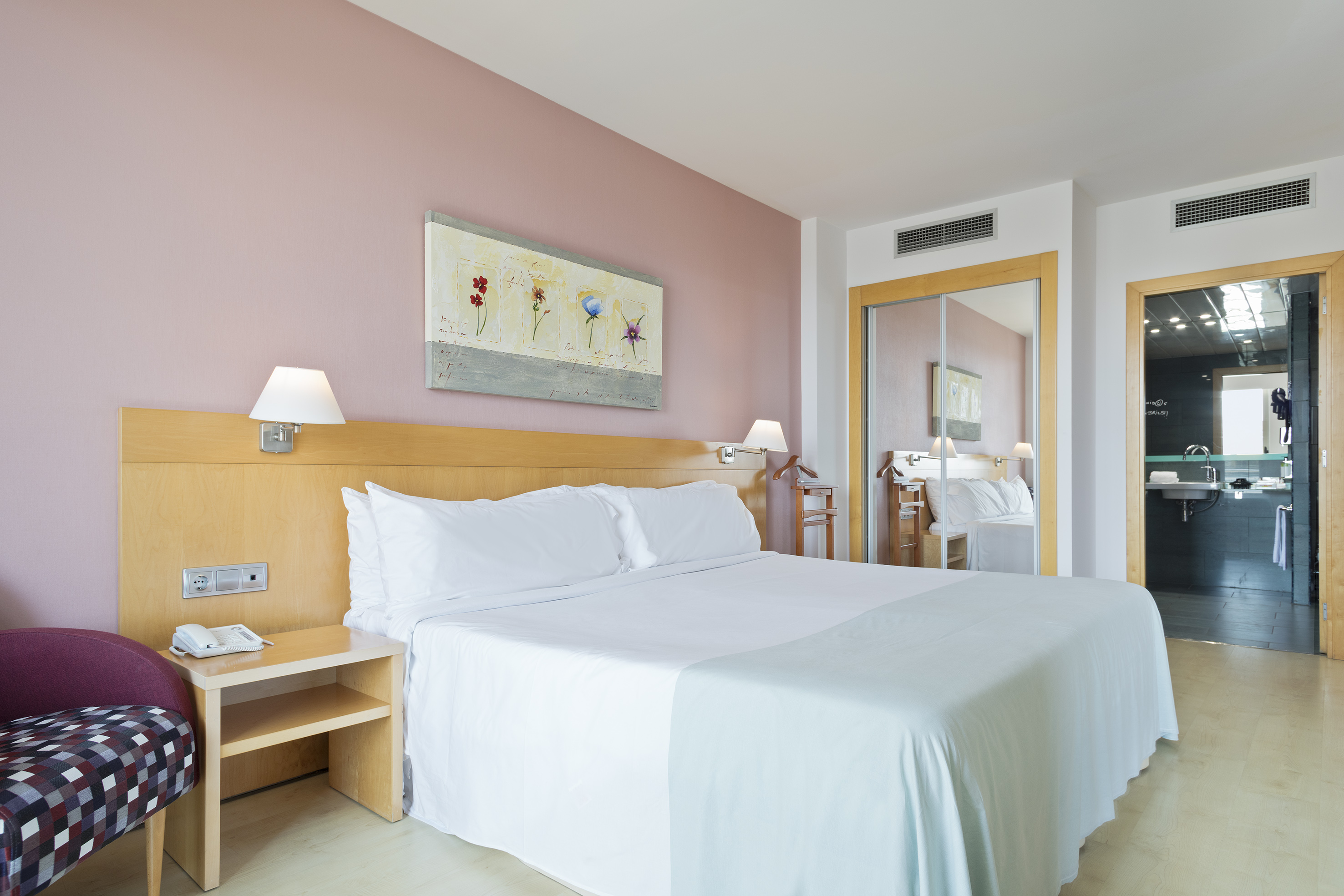 Modern hotel bedroom with queen bed, pink wall, artwork, and visible bathroom.