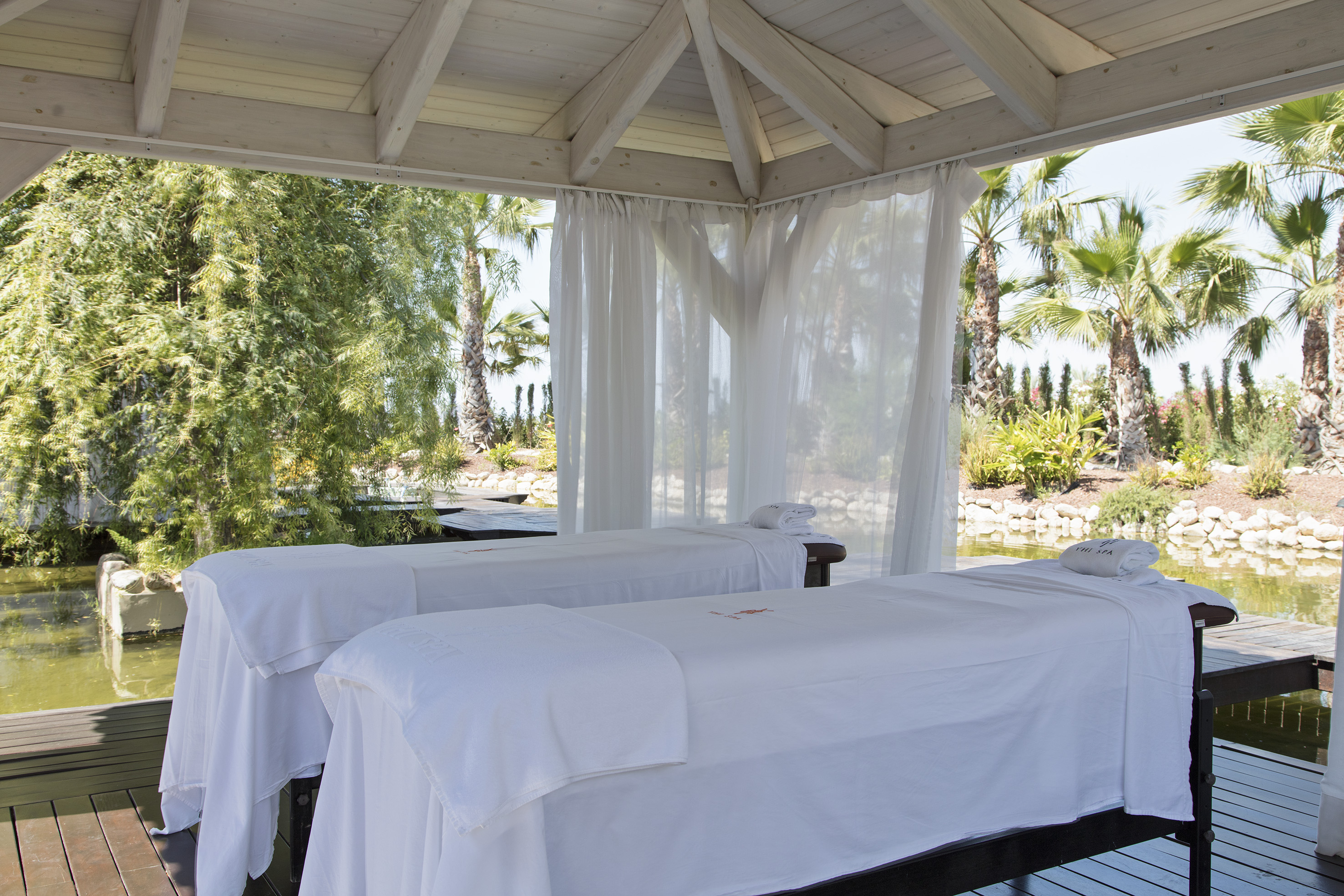 Tropical outdoor spa gazebo with two massage beds, pond, and palm trees.
