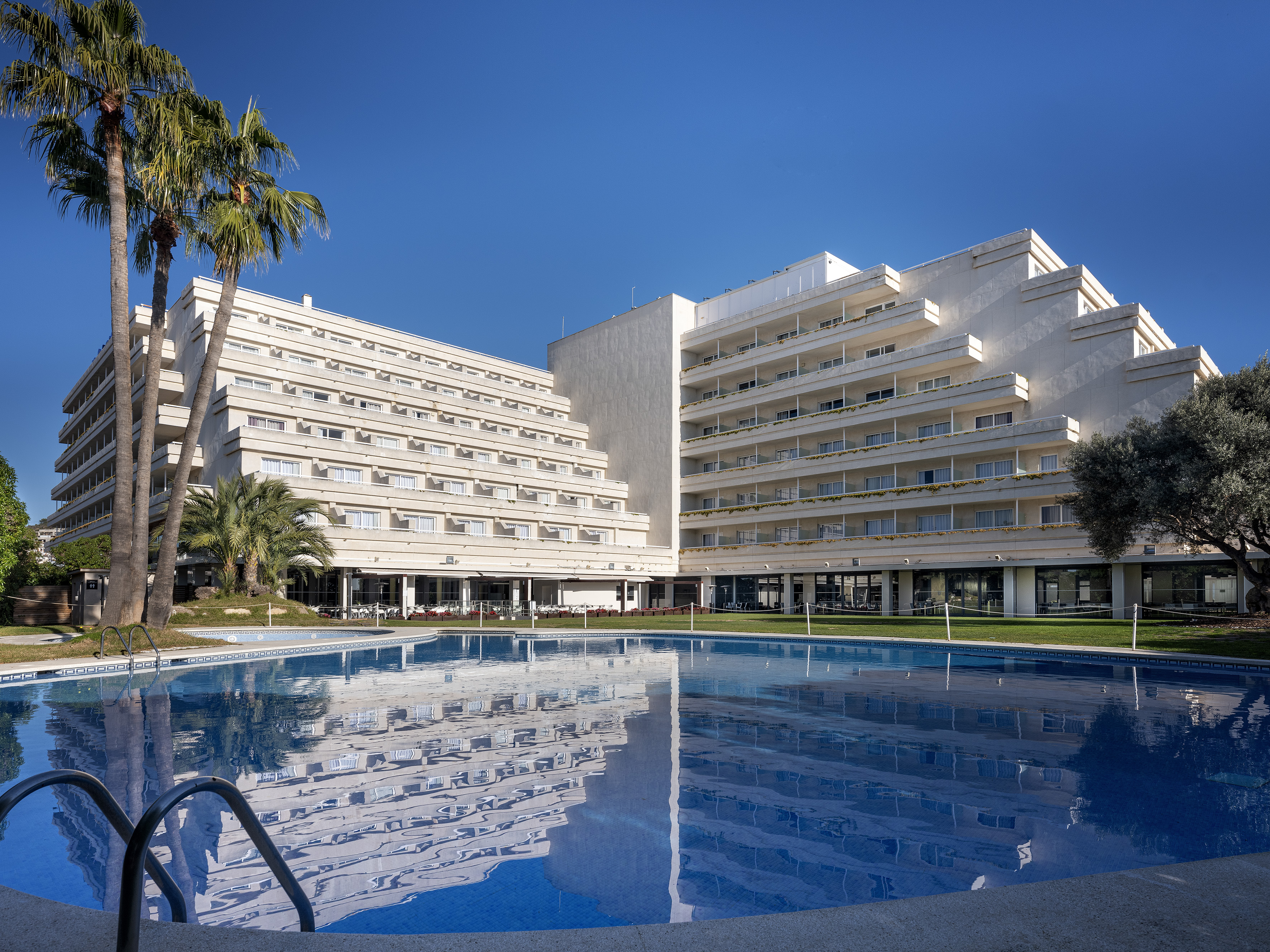 White hotel resort with terraced buildings, outdoor pool, and palm trees.
