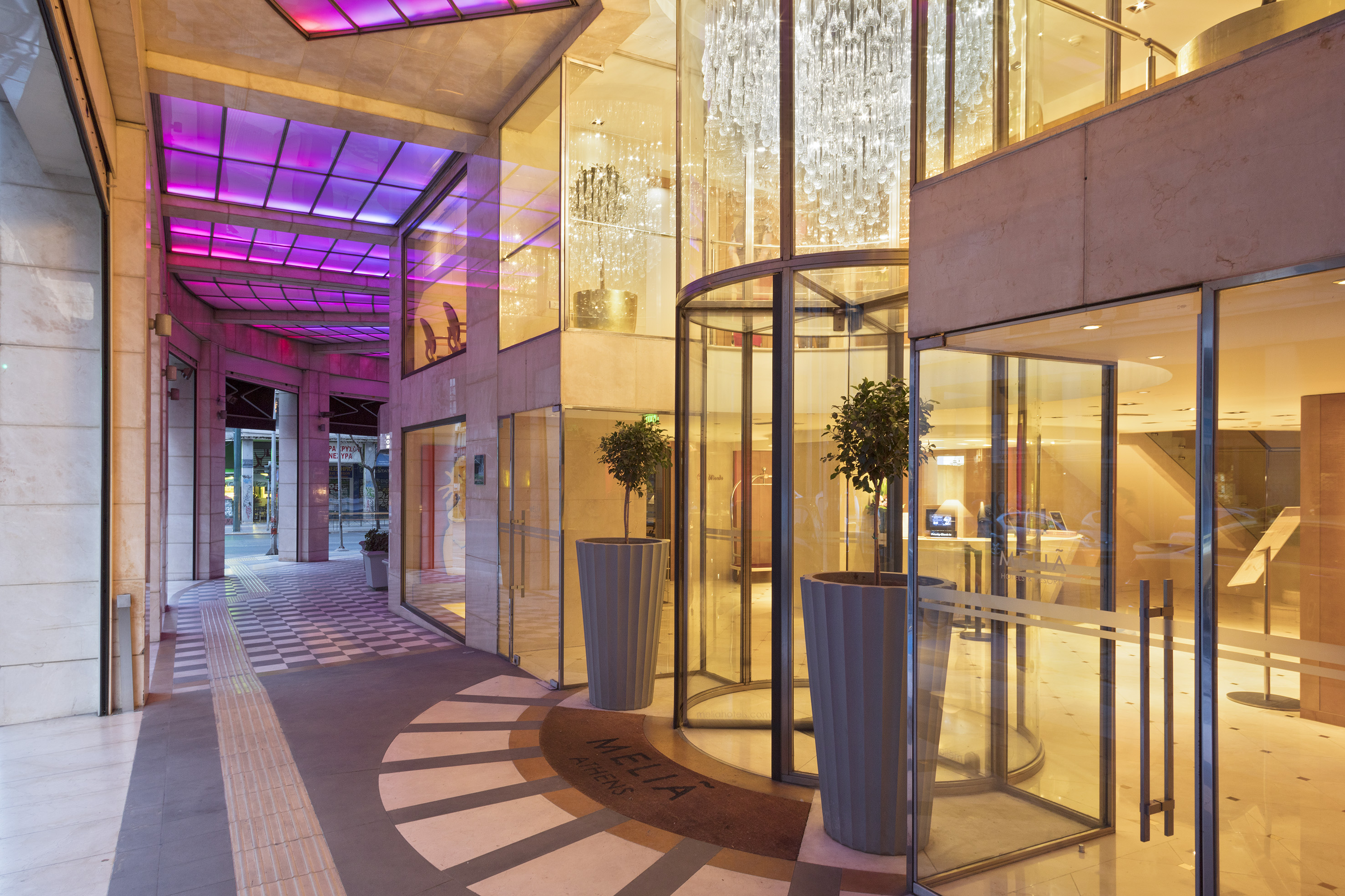 Meliá Athens hotel entrance with glass doors, chandeliers, and pink-lit canopy.