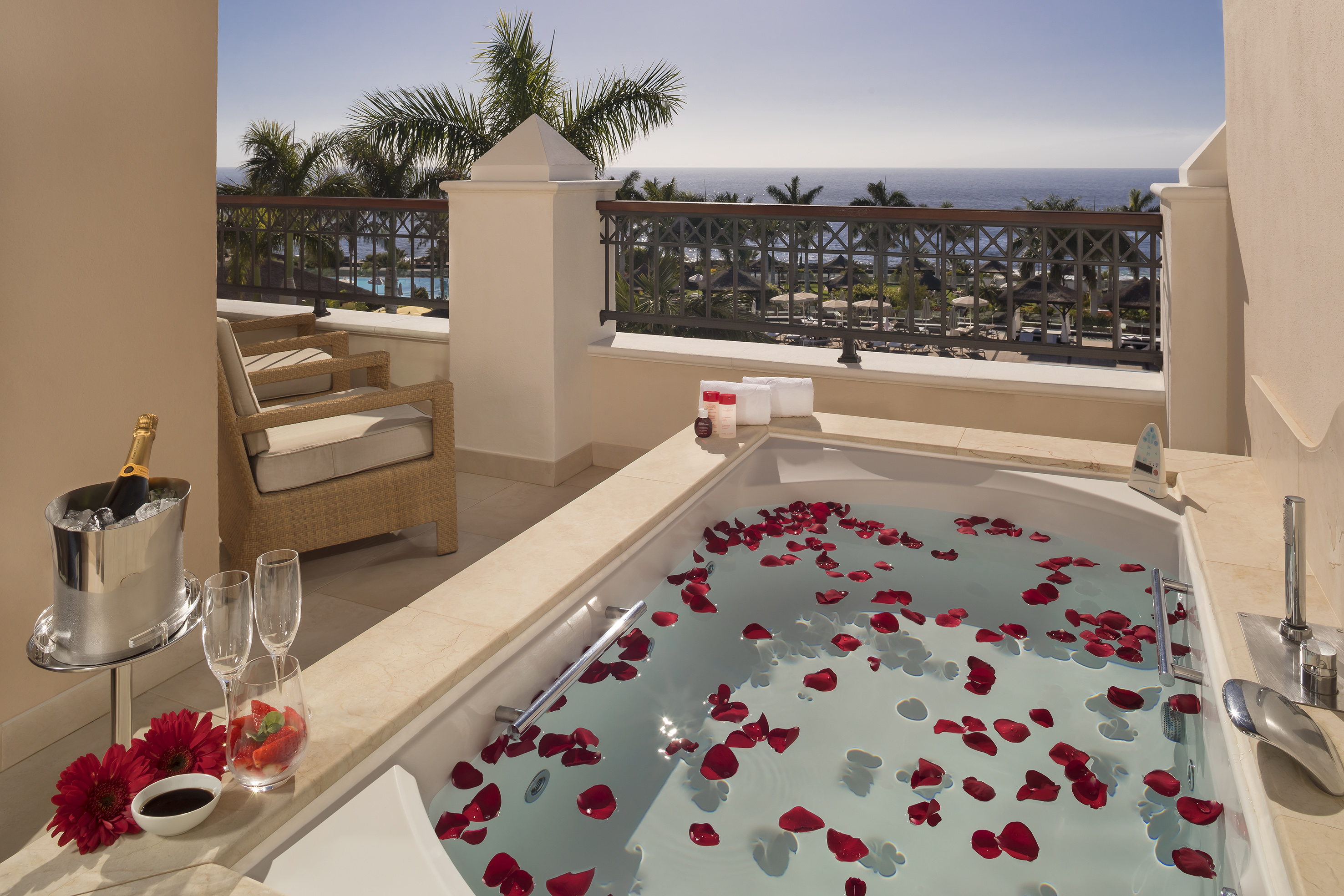 Romantic balcony jacuzzi with ocean view, rose petals, and champagne setup.