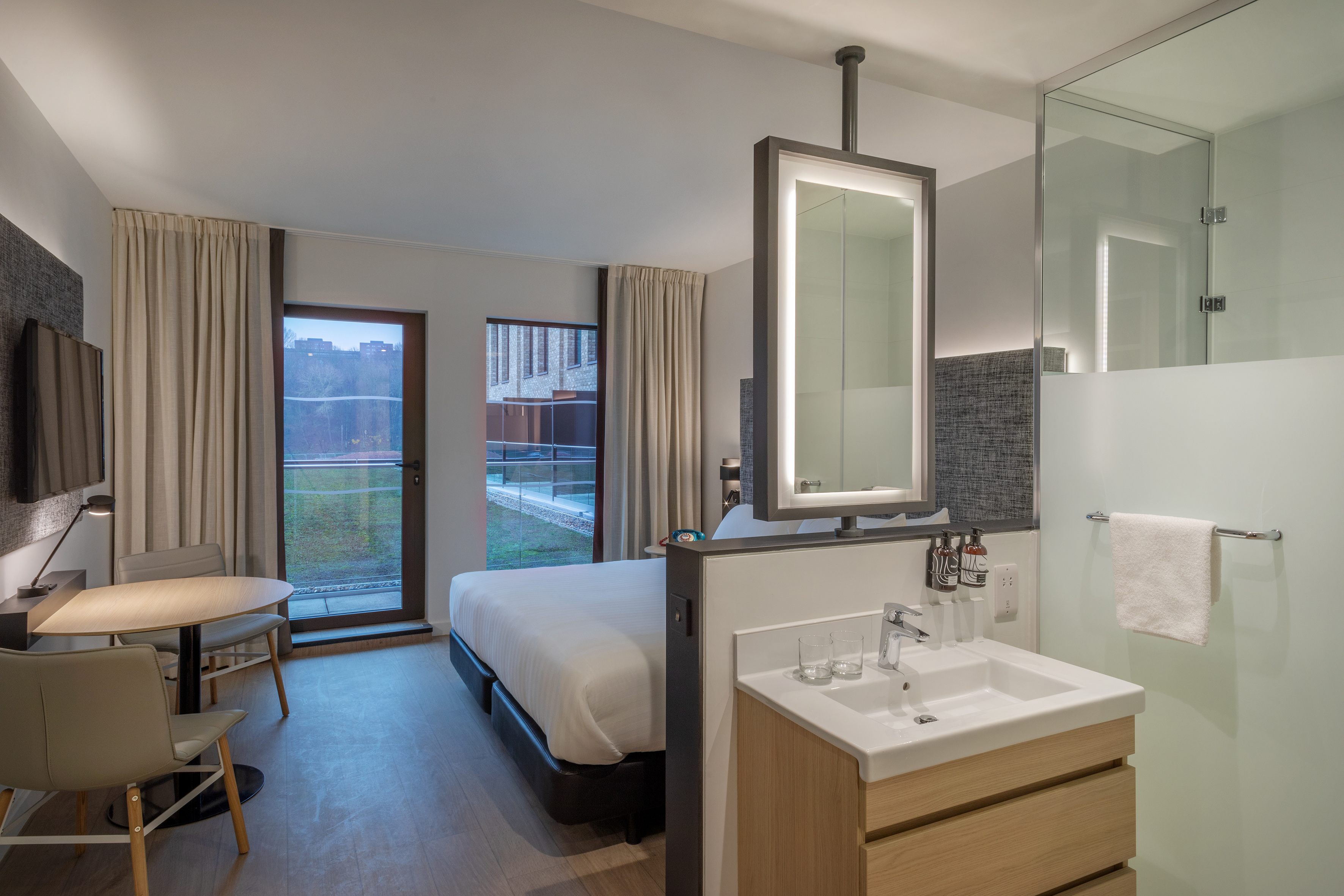 Modern hotel room interior with bed, small desk, and integrated bathroom vanity.