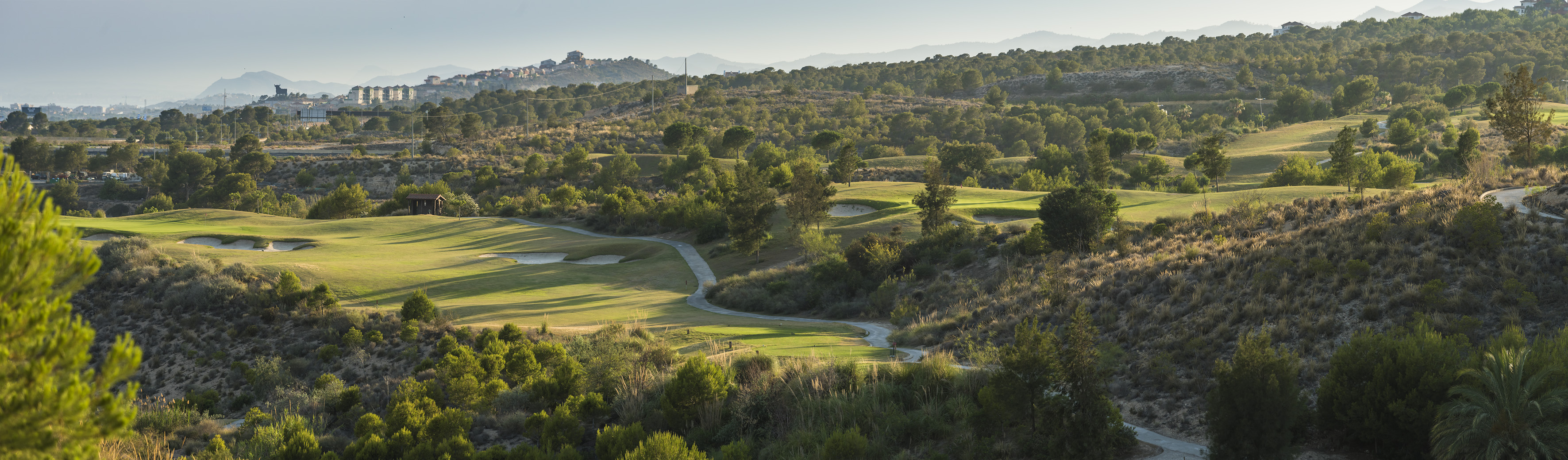 Panoramic hilly golf course with green fairways, bunkers, and winding path.