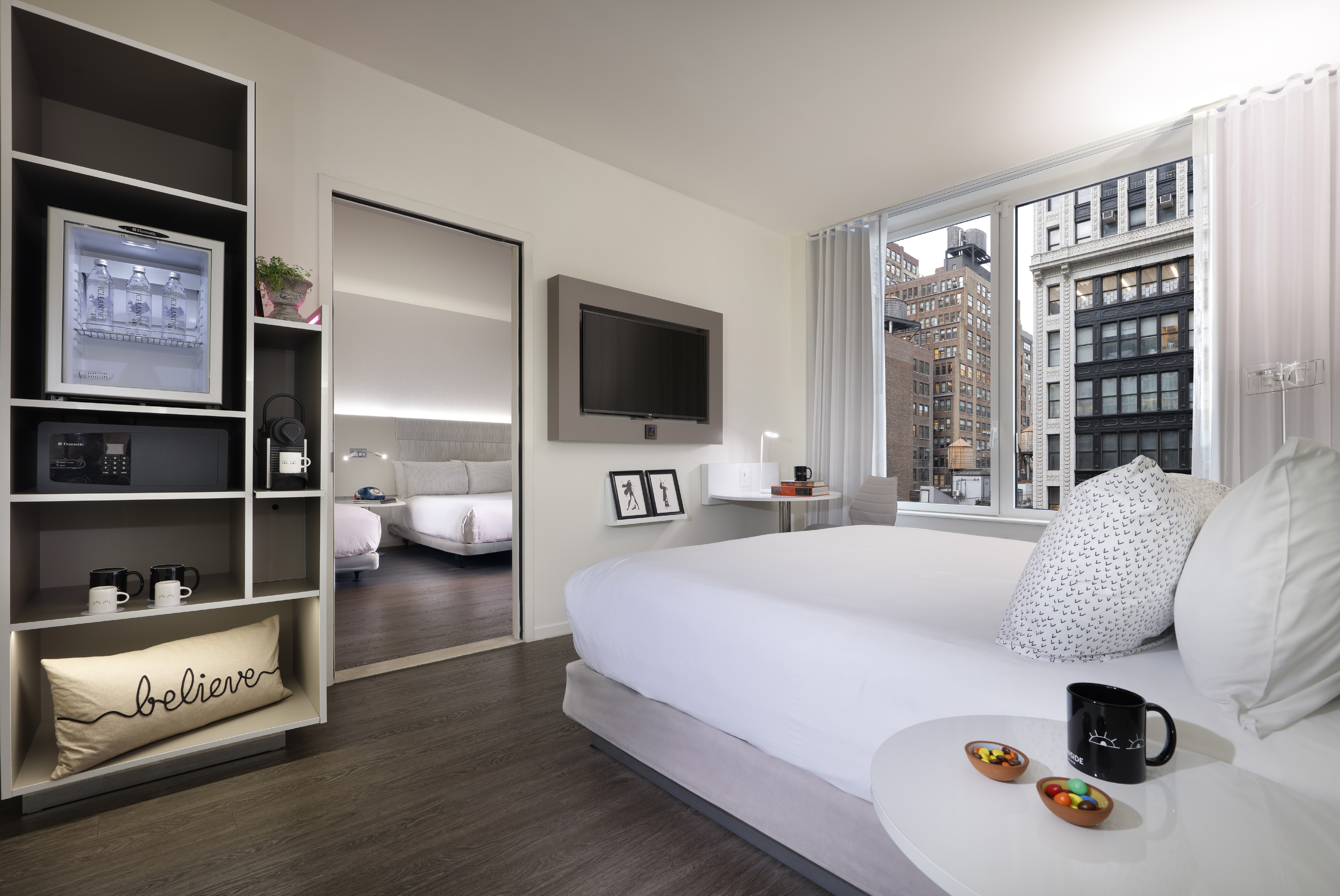 Modern hotel room with city view, king bed, TV, minibar, and adjacent bedroom.