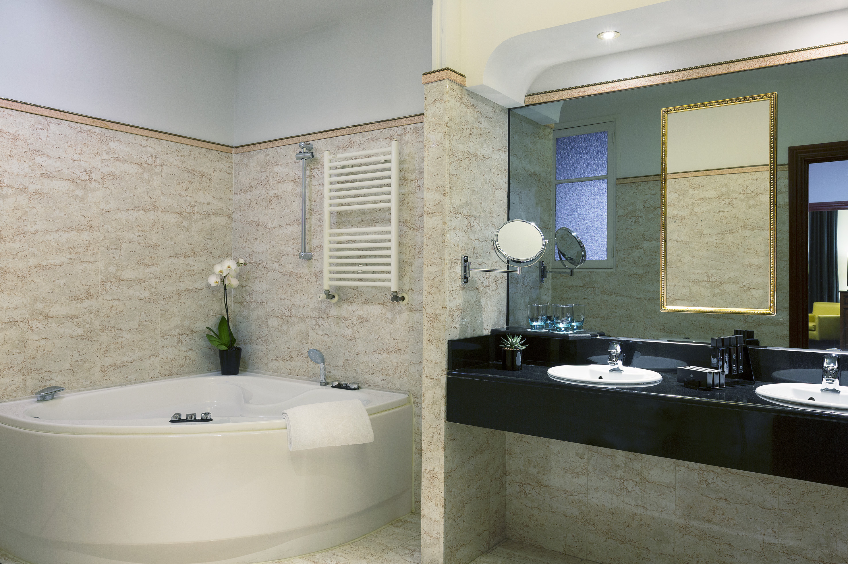 Hotel bathroom with corner tub, marble walls, and black double vanity.