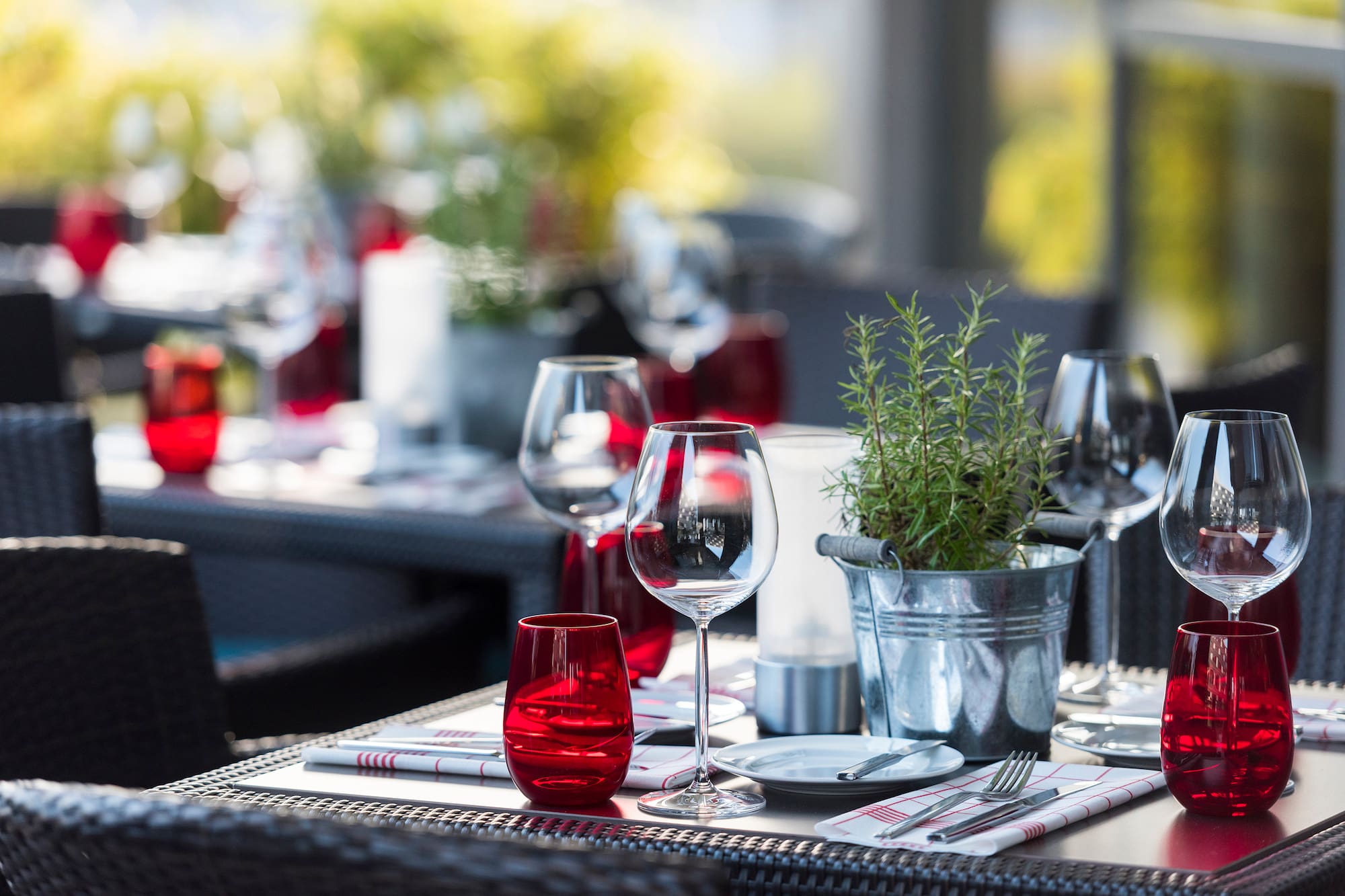 Outdoor restaurant table set with wine glasses, red tumblers, and rosemary plant.
