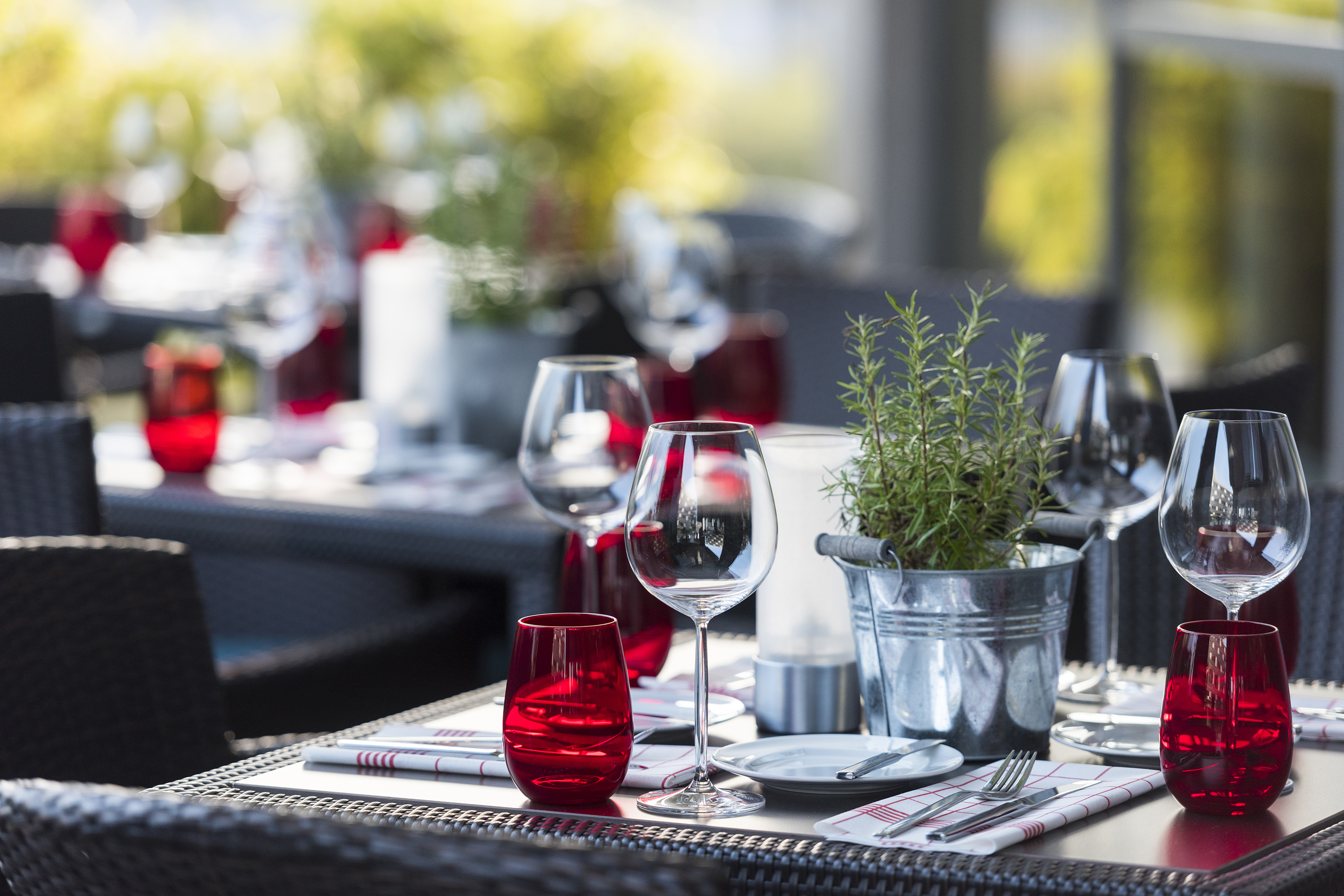 Outdoor restaurant table set with wine glasses, red tumblers, and rosemary plant.