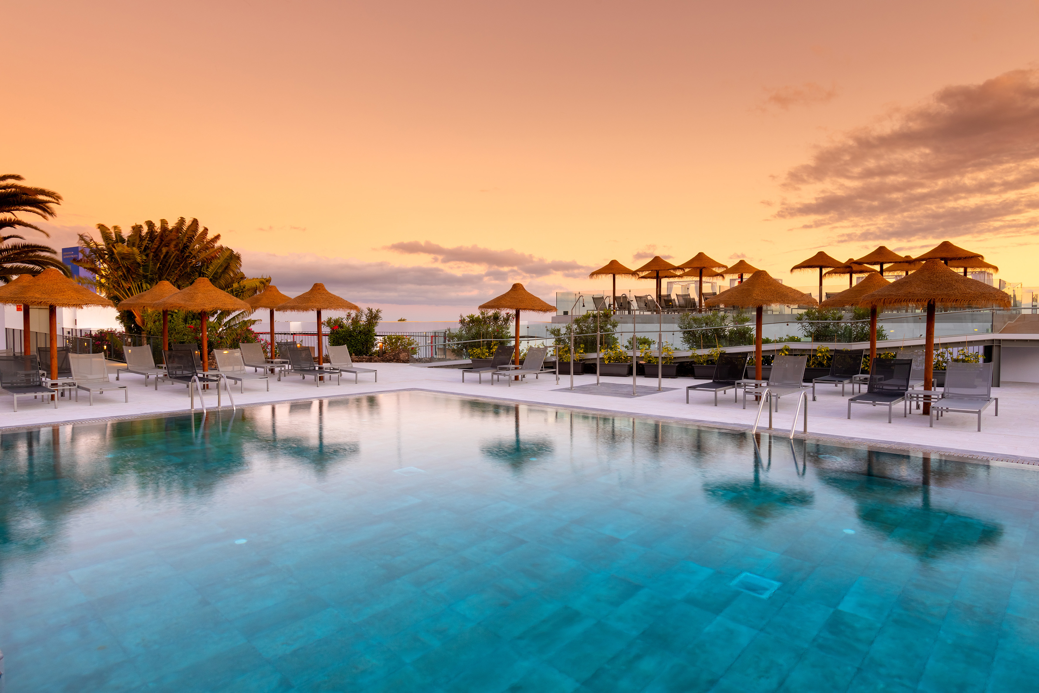 Tropical resort pool with lounge chairs, thatch umbrellas, and sunset sky.