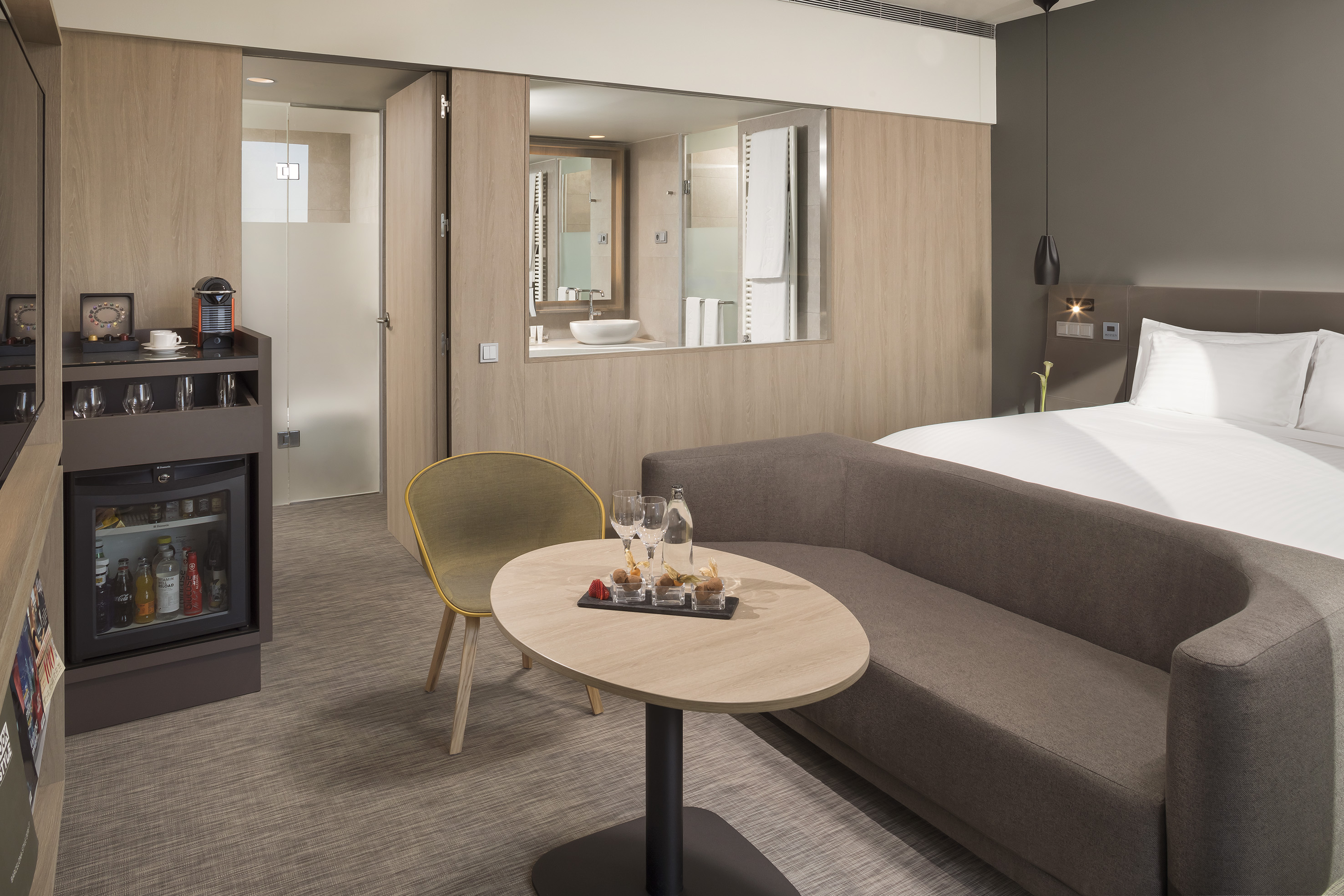Modern hotel room with comfortable bed, sofa, minibar, and integrated bathroom.