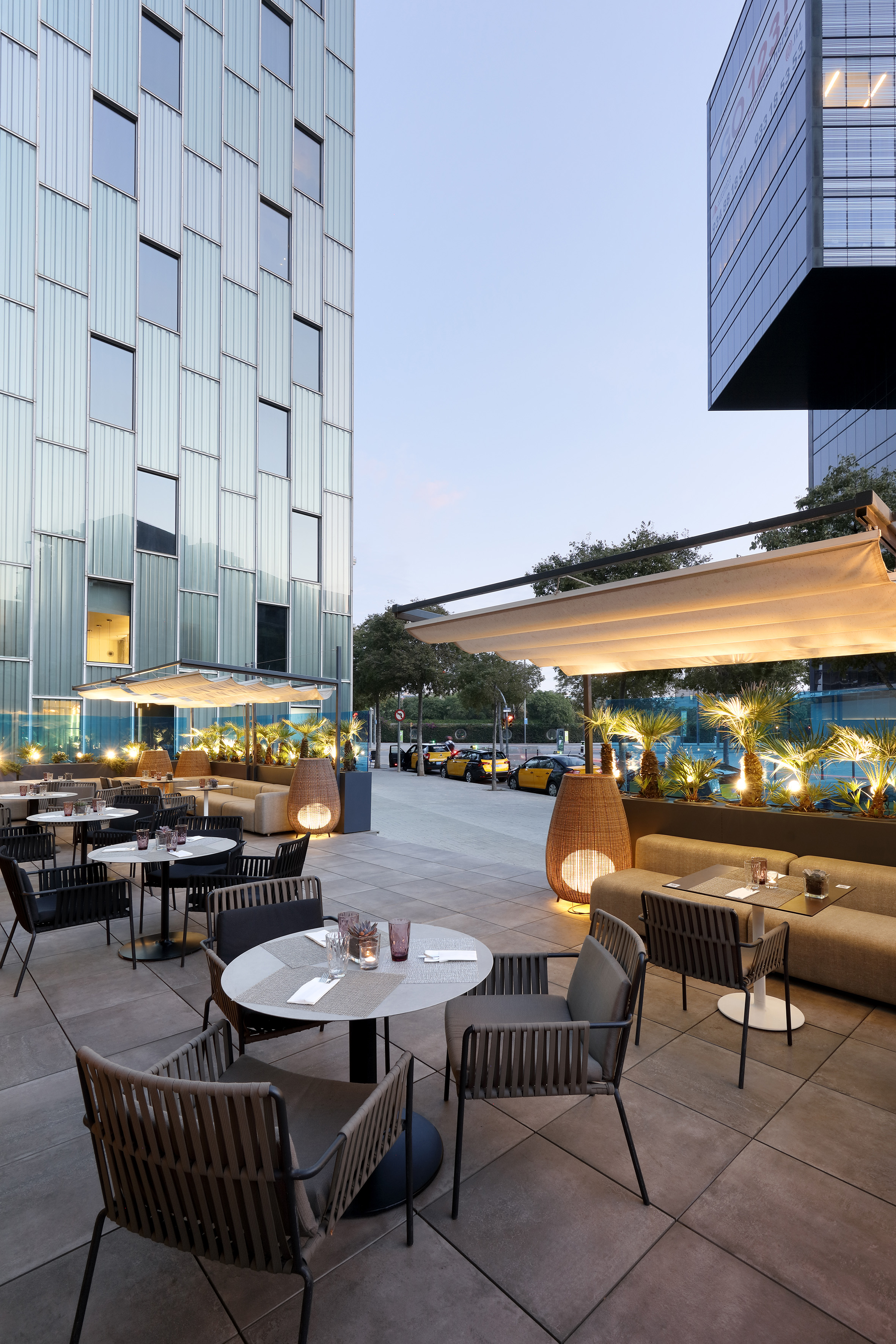 Modern outdoor restaurant terrace with evening lighting, seating, and glass buildings.