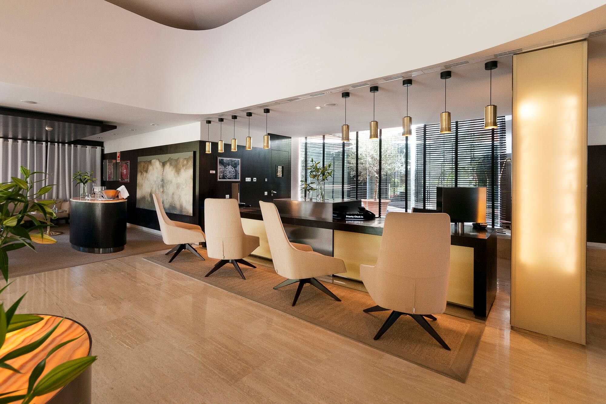 Modern hotel lobby with long reception desk, beige chairs, and gold pendant lights.