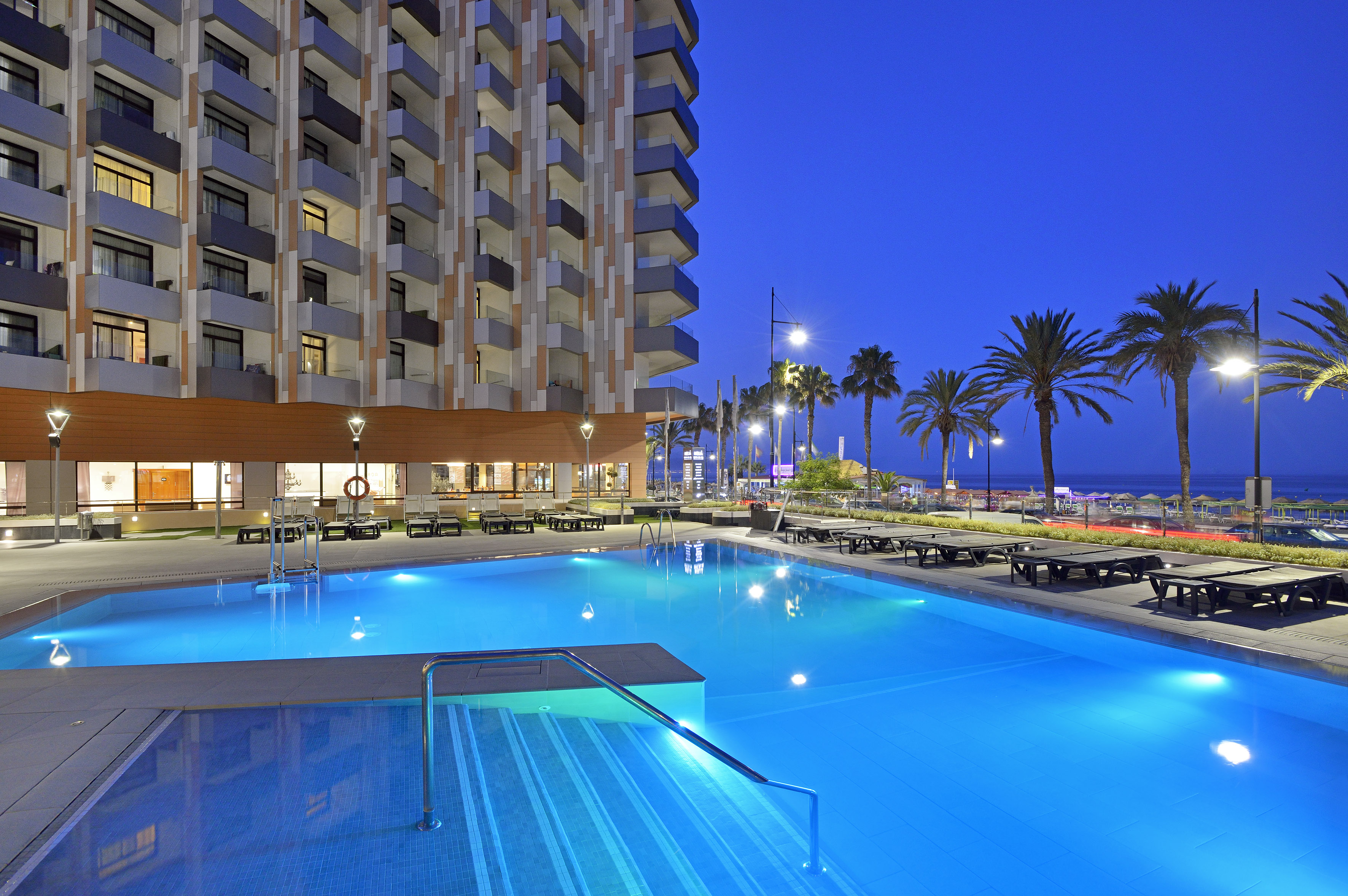 Modern hotel with illuminated pool, palm trees, and ocean view at dusk.