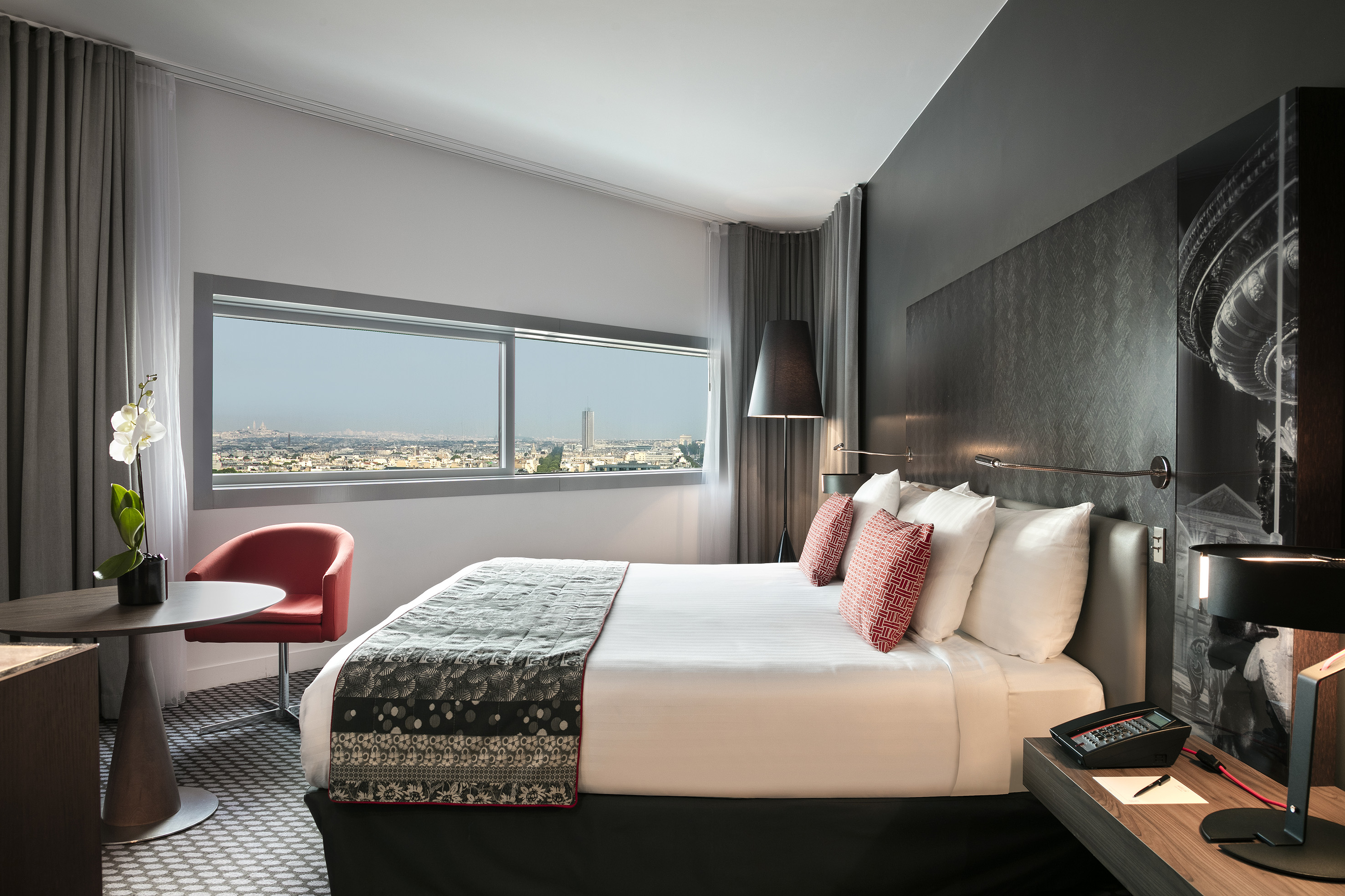 Sleek hotel room with city skyline view, large bed, and red accent chair.
