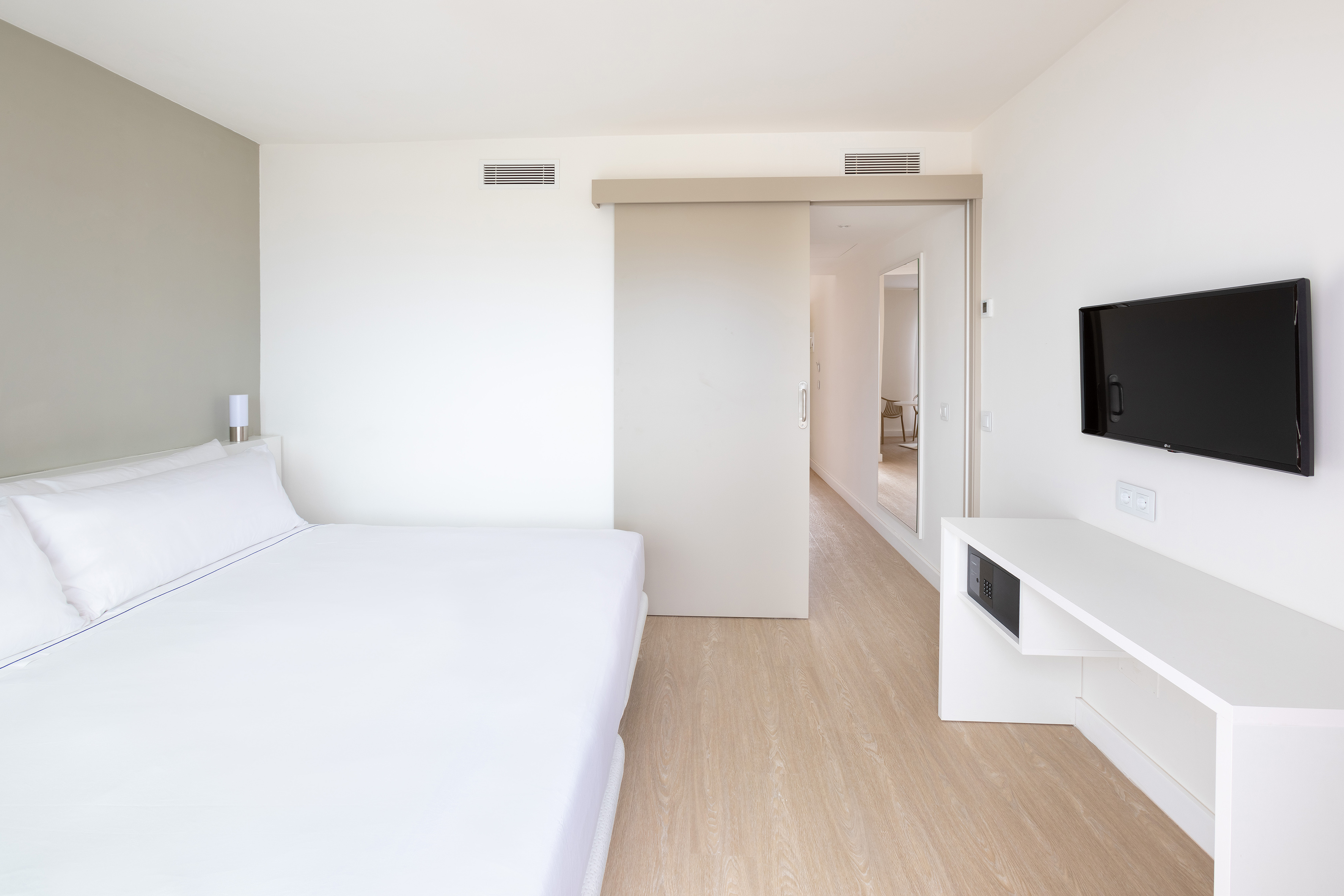 Bright, modern hotel room with white bed, TV, wood floor, and sliding door.