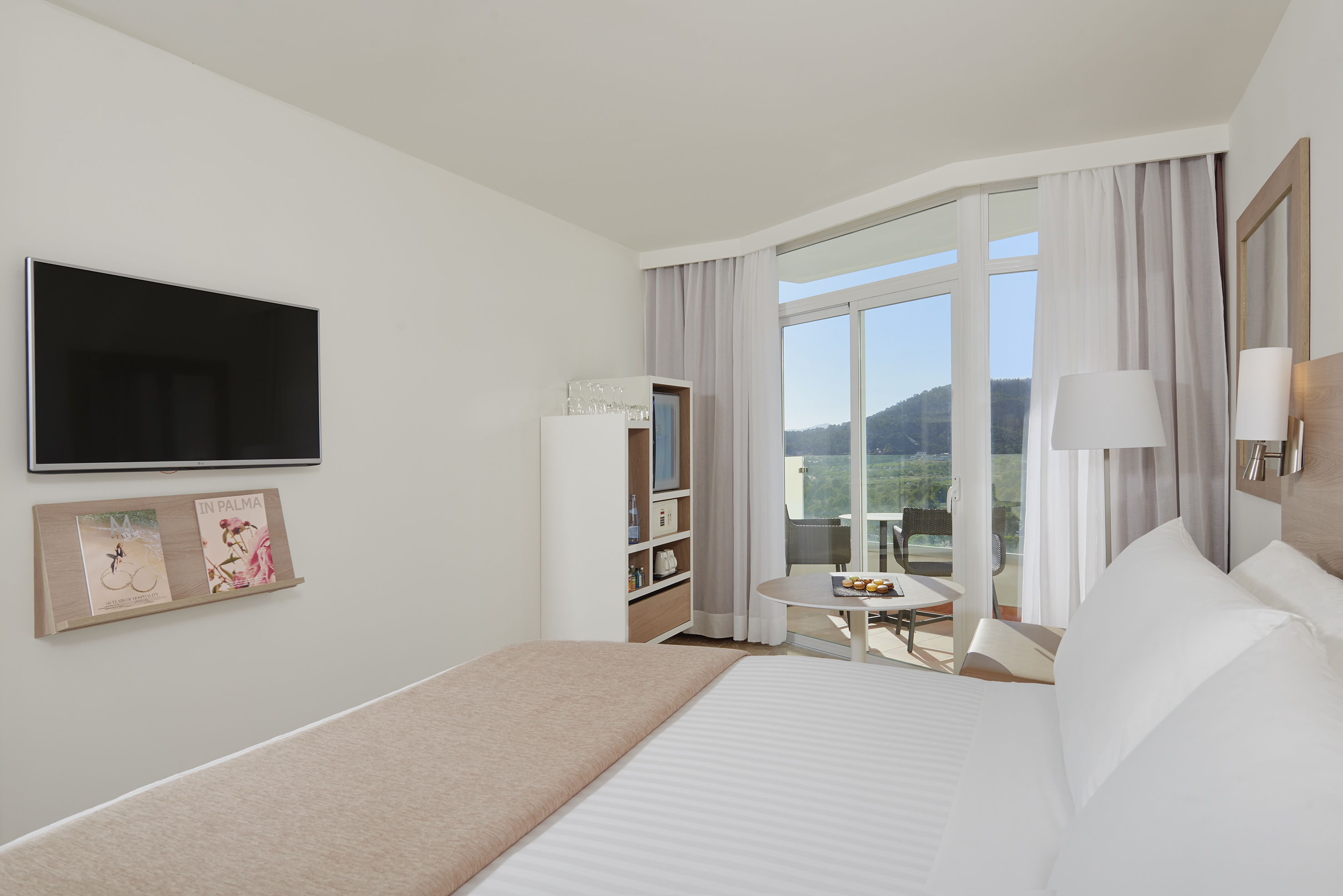 Modern hotel room with bed, TV, mini-bar, and balcony with mountain view.